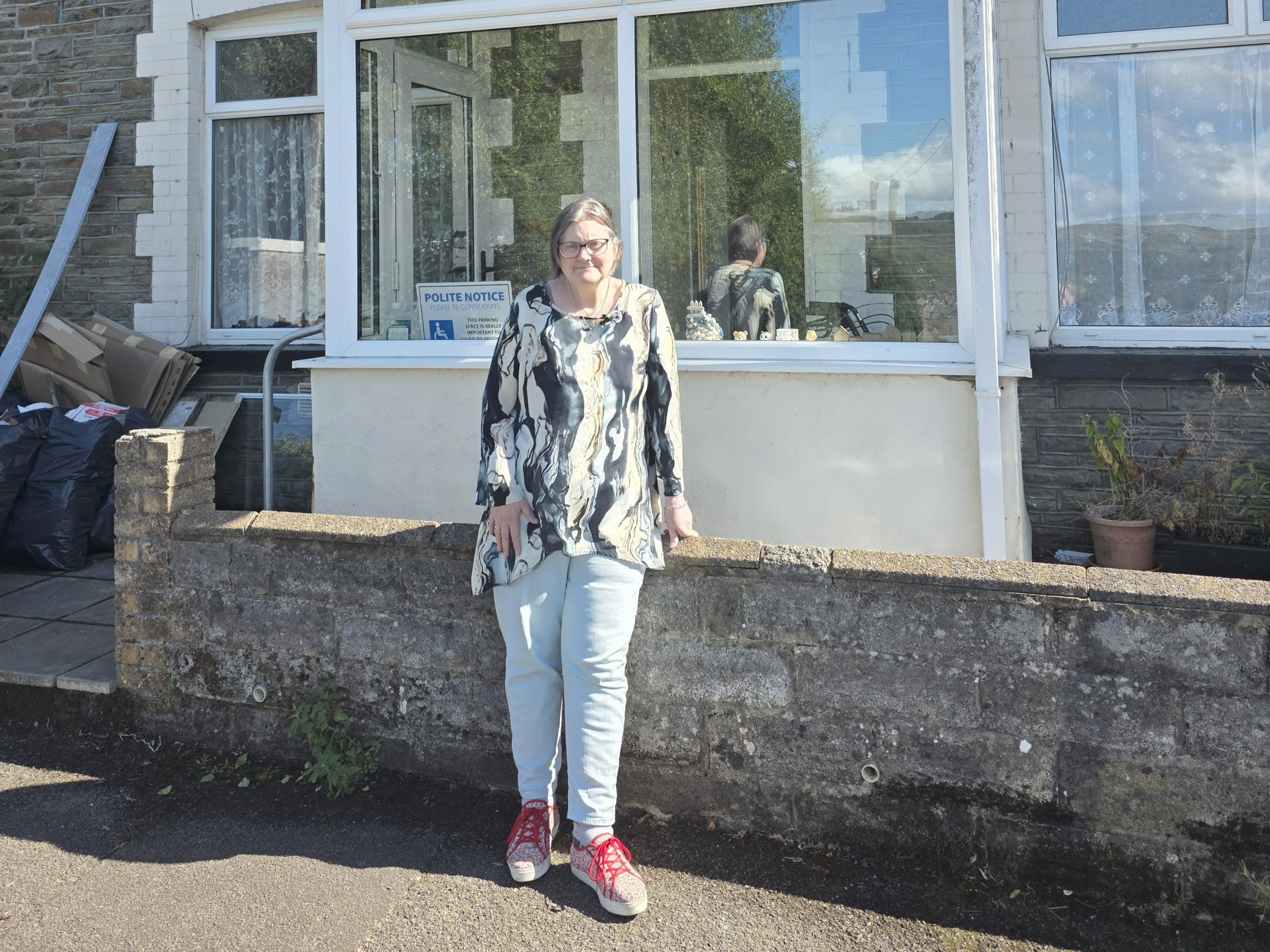 Trivallis Housing Landlord Wales A person with light-colored hair, glasses, a patterned top, light jeans, and red shoes stands in front of a low stone wall outside a building with large windows. Some bags and a potted plant are visible nearby.