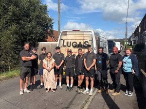 Trivallis Housing Landlord Wales A group of eleven people stand on a residential street in front of a van with the word “LUCAS” on it. Most are wearing black clothing. The weather is sunny with some clouds in the sky.