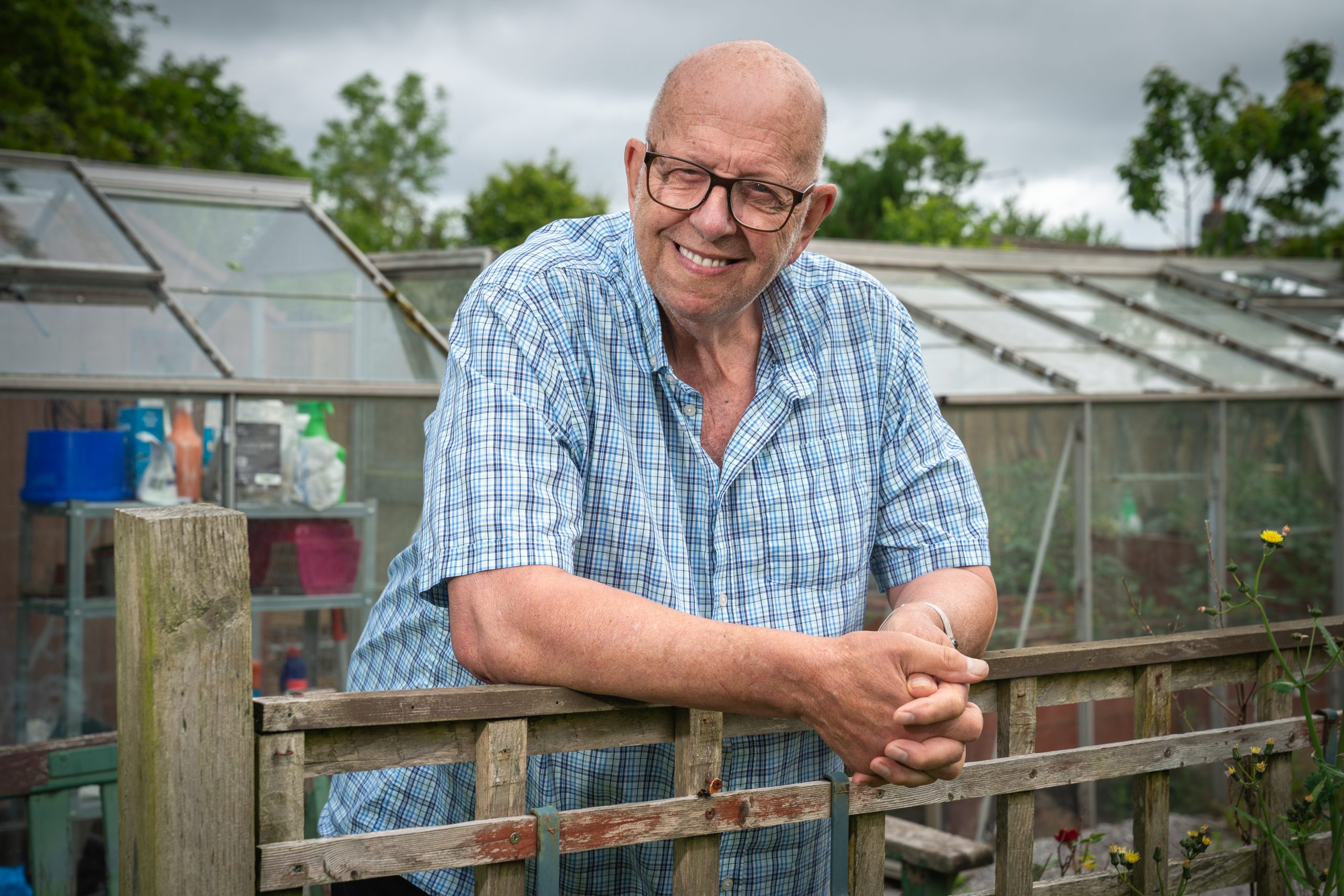 Trivallis Housing Landlord Wales An older man with glasses and a bald head, wearing a blue plaid shirt, leans on a wooden fence in front of a greenhouse and garden on a cloudy day.