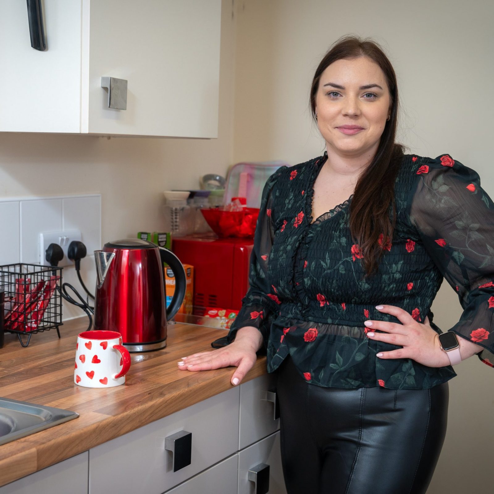 Trivallis Housing Landlord Wales A woman with long brown hair stands in a kitchen beside a wooden countertop with a red kettle, a mug with red hearts, and various kitchen items. She is wearing a black floral blouse and black pants.