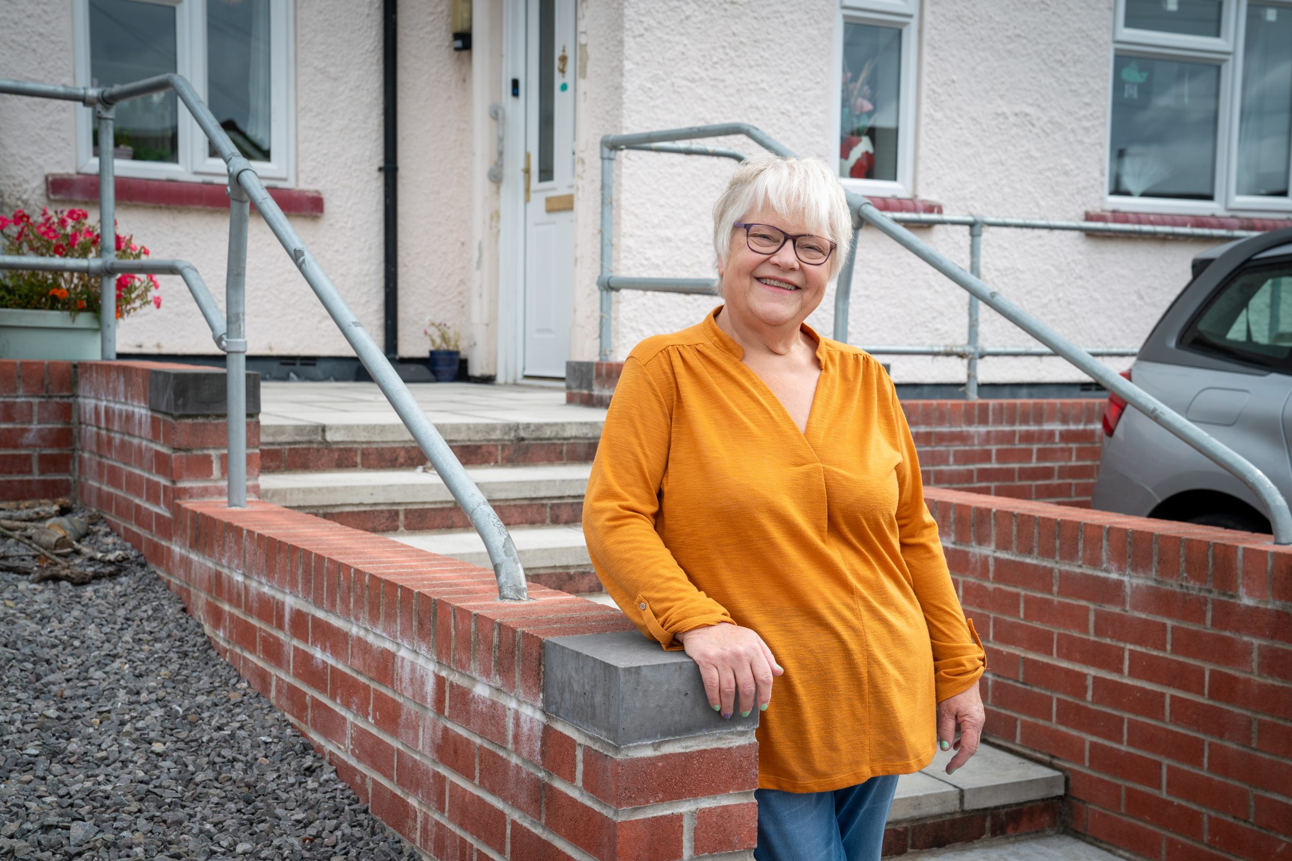 Trivallis Housing Landlord Wales An older woman with short white hair and glasses, wearing a mustard yellow top, stands and smiles by a brick wall and metal handrail outside a house with a car parked nearby.
