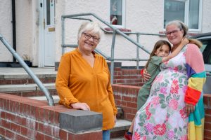 Trivallis Housing Landlord Wales Three people pose outside a building: an older woman in an orange shirt stands by a brick railing, while a woman in a floral dress and rainbow cardigan hugs a young child on the steps behind her.