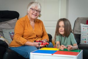 Trivallis Housing Landlord Wales An older woman with short gray hair and glasses sits on a couch next to a young child with long brown hair. They are playing with a toy car and a colorful building block table in a living room.
