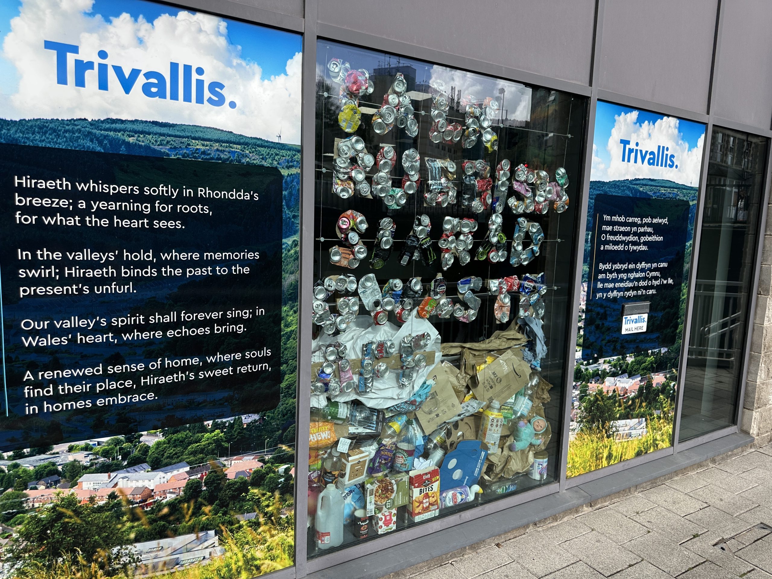Trivallis Housing Landlord Wales A storefront window display shows a large pile of crushed cans and plastic bottles, with informational posters about recycling and the environment from Trivialis visible on the glass.