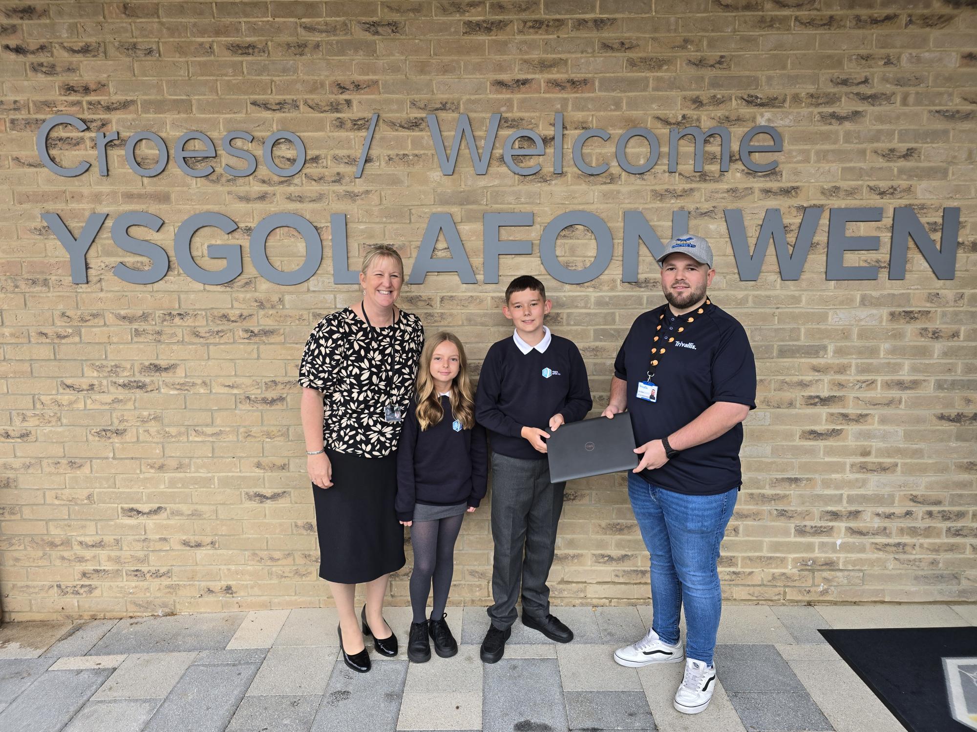 Trivallis Housing Landlord Wales Four people stand in front of a brick wall with the sign "Croeso / Welcome YSGOL AFON WEN." One person hands a laptop to a student while the others stand beside them, all posing for the photo.