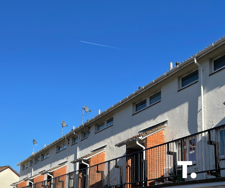 Trivallis Housing Landlord Wales A clear blue sky with a white contrail overhead, above a row of modern townhouses with brick and white facades, railings, and satellite dishes. A white letter "T." is in the lower right corner.