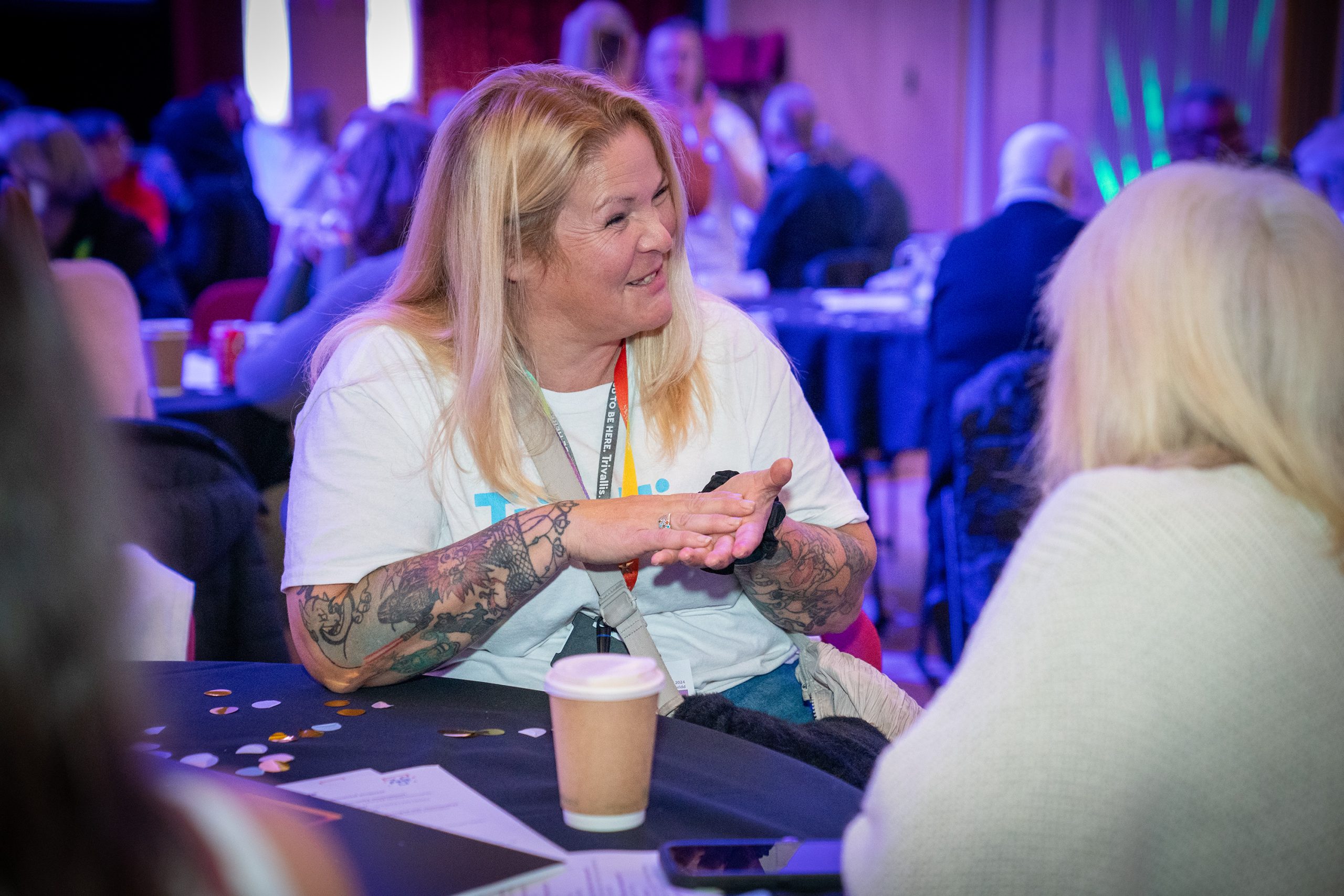 Trivallis Housing Landlord Wales A woman with long blonde hair and tattoos on her arms sits at a round table, talking to another person. There are papers, a paper coffee cup, and scattered confetti on the table. People are in the background.