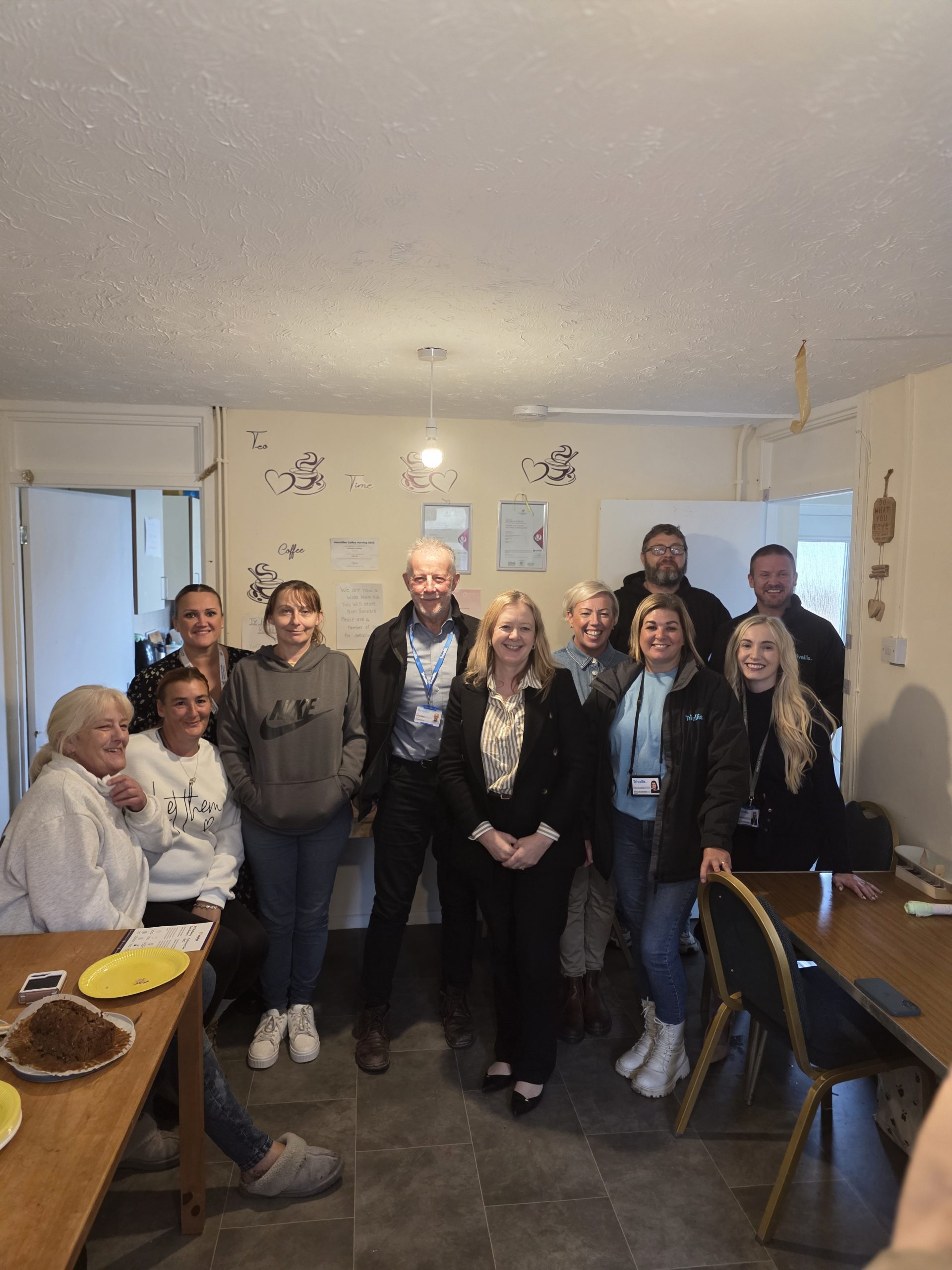 Trivallis Housing Landlord Wales A group of eleven people, some seated and some standing, pose and smile for a photo in a kitchen or dining area with a table, chairs, and wall decorations visible in the background.