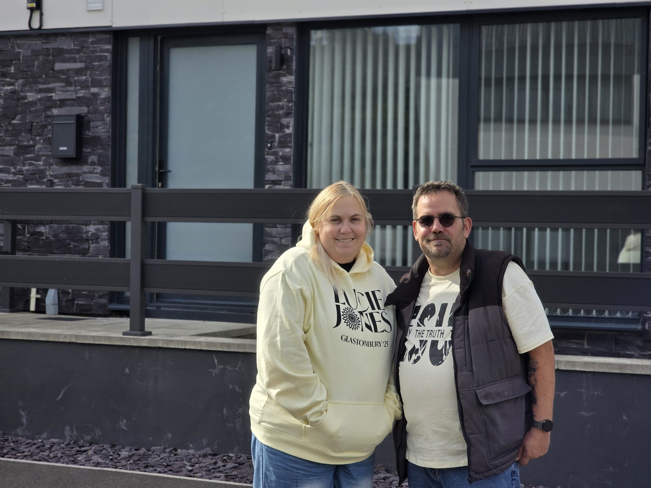 Trivallis Housing Landlord Wales Two people stand side by side outdoors in front of a modern building with large windows and a black fence. Both are wearing light-colored tops and looking at the camera.