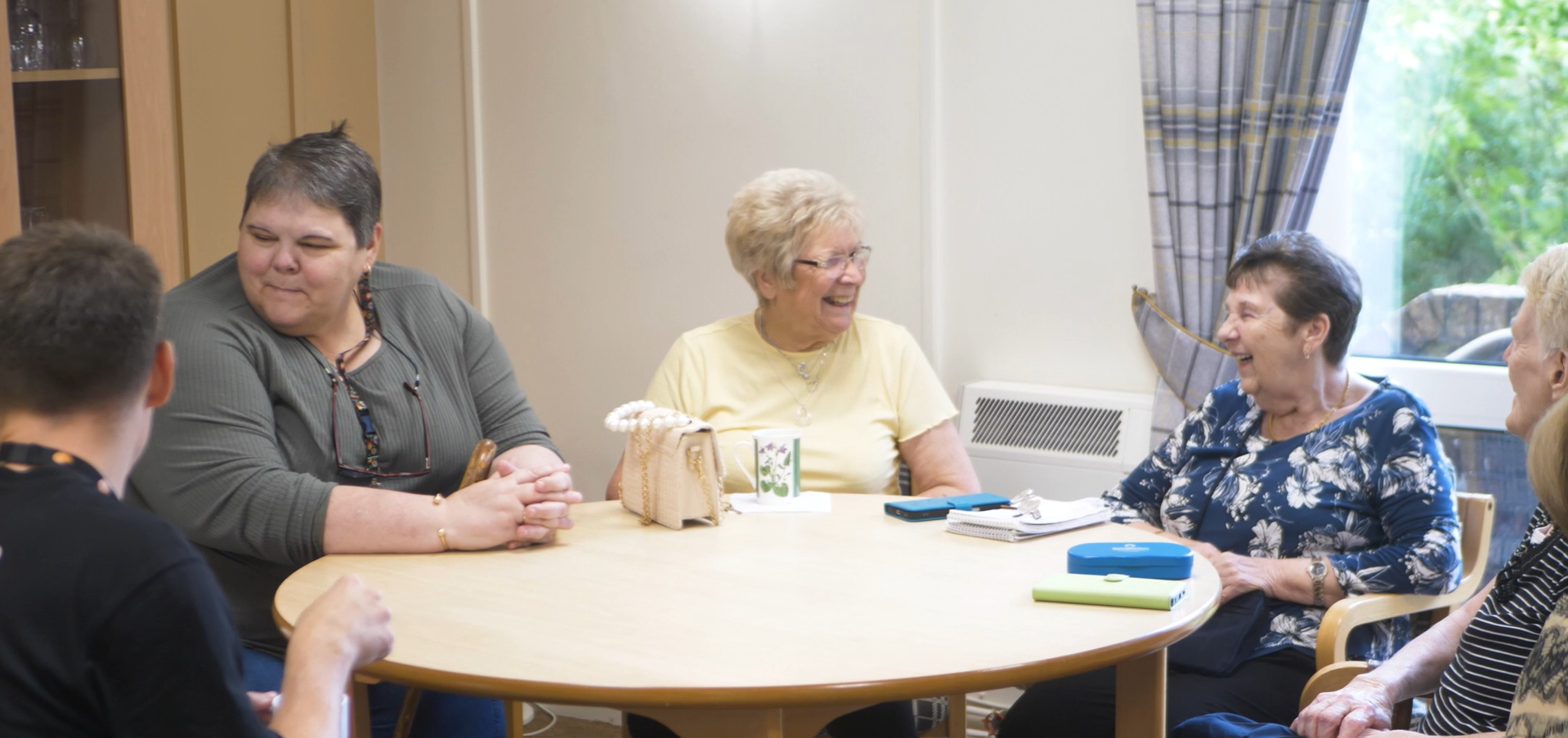 Trivallis Housing Landlord Wales Four older adults sit around a round wooden table, engaged in conversation and smiling. Notebooks, a cup, and a bag are on the table. The setting is indoors with a window in the background.