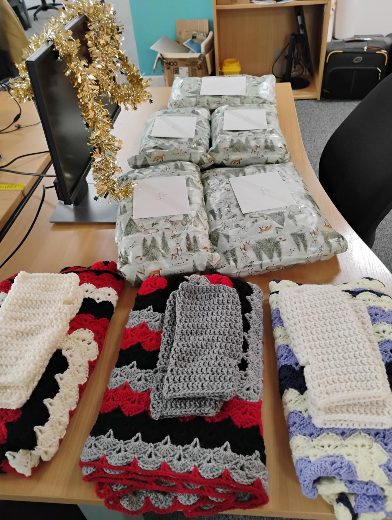 Trivallis Housing Landlord Wales Four folded crocheted blankets with matching washcloths are laid out on a desk, in front of five gift-wrapped packages. The desk has a computer monitor decorated with gold tinsel and office furniture in the background.