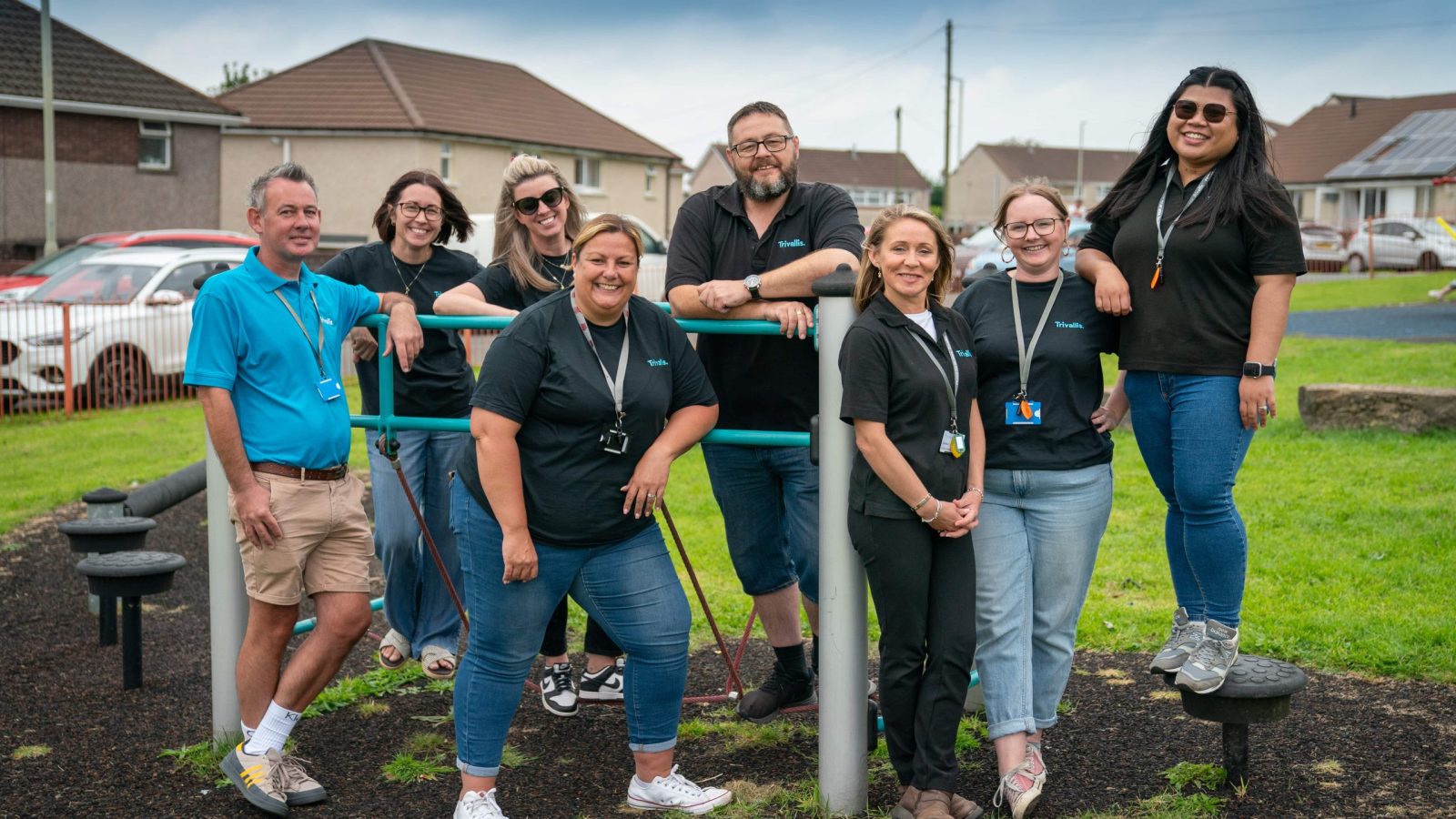 Trivallis Housing Landlord Wales A group of eight adults poses together outdoors at a playground. Most are wearing black shirts and name badges. The background shows houses, grass, parked cars, and playground equipment.