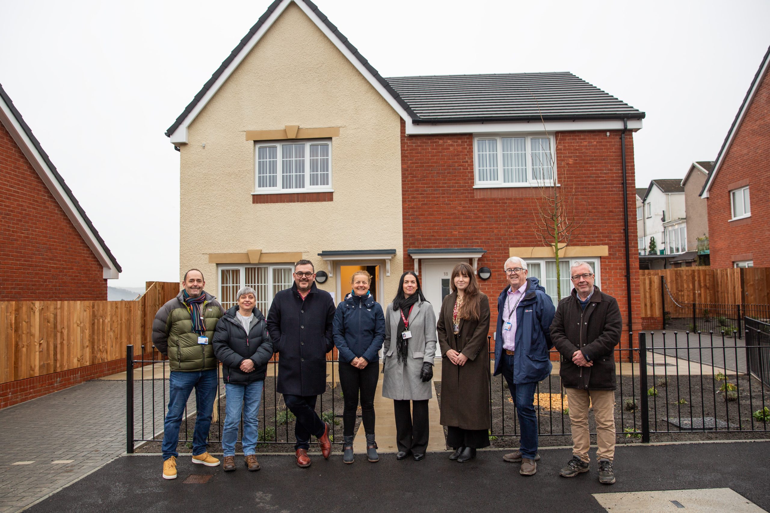 Trivallis Housing Landlord Wales Eight people stand in a row outside a modern two-story house with a beige and red brick exterior. The group stands on a paved driveway with a black fence in front of the house. It appears cloudy.