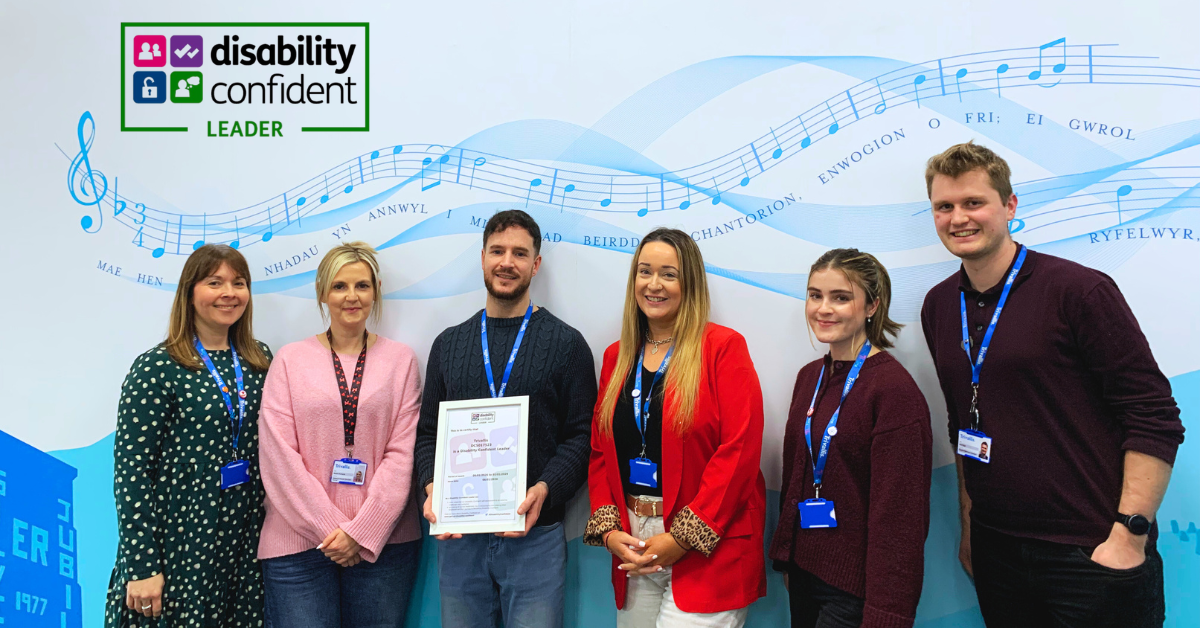 Trivallis Housing Landlord Wales Six people stand in a row, some smiling, in front of a wall with musical notes and blue design. One person holds a framed certificate. A "Disability Confident Leader" logo appears in the top left corner.