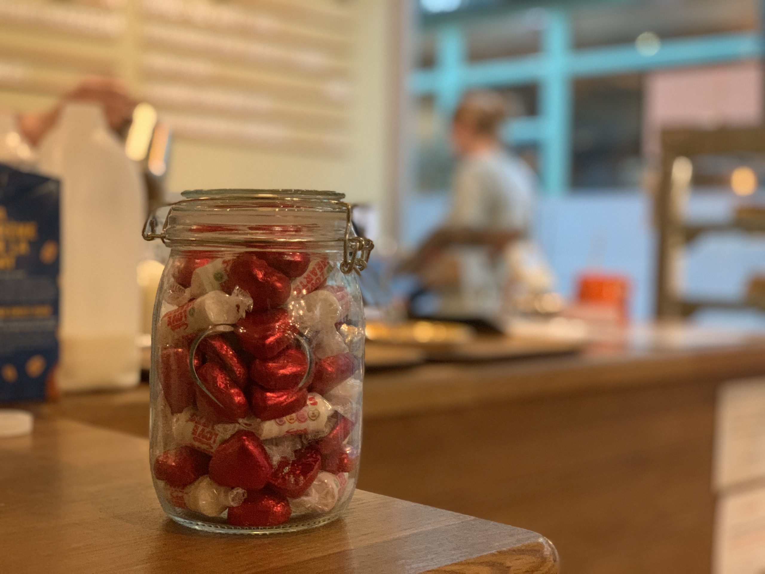 Trivallis Housing Landlord Wales A glass jar filled with red and white wrapped candies sits on a wooden counter in what appears to be a cafe or bakery, with people and shelves blurred in the background.