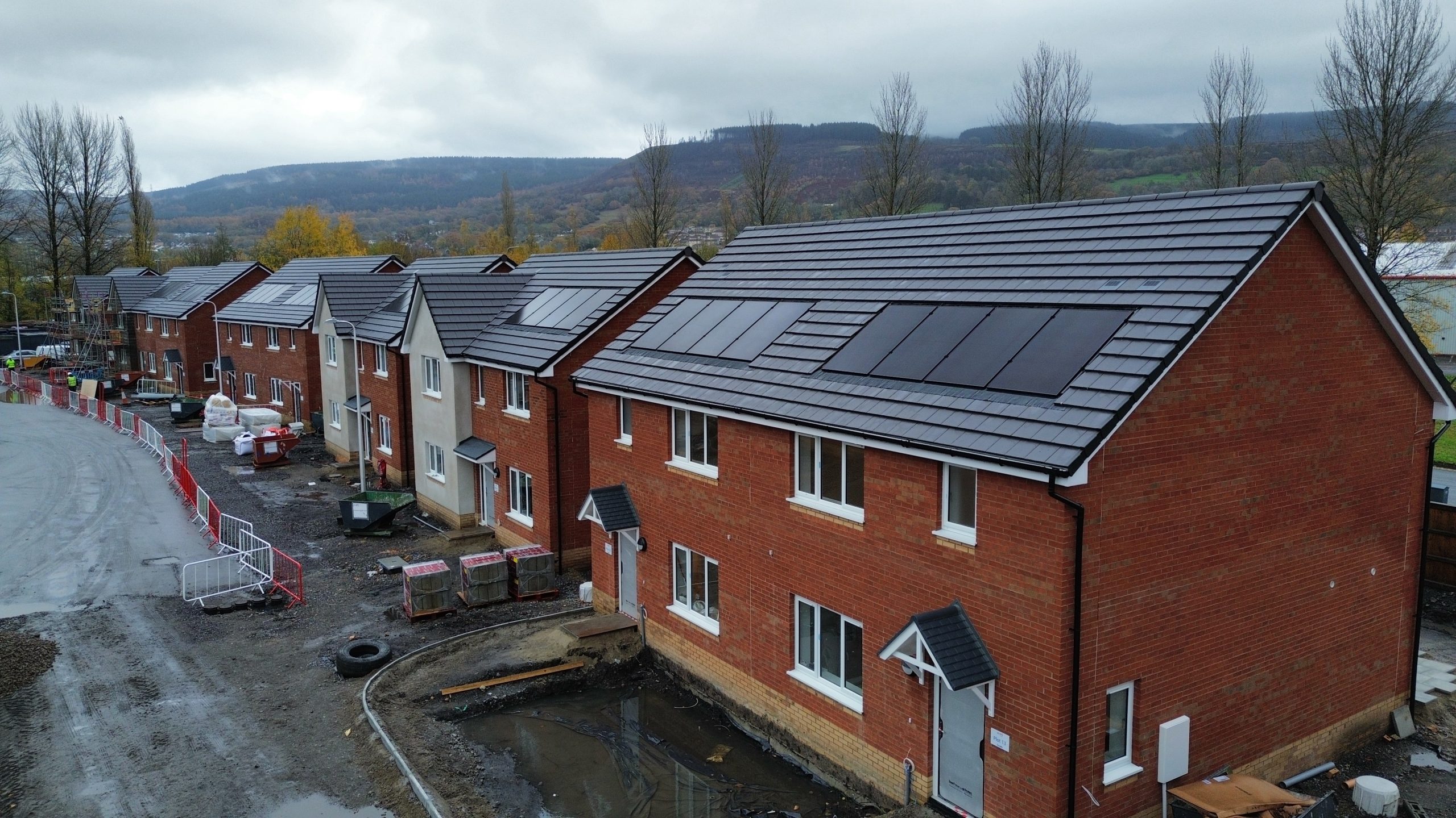 Trivallis Housing Landlord Wales A row of new red-brick houses with grey roofs, some fitted with solar panels. The ground outside appears muddy and unfinished, with construction materials and fencing present. Trees and hills are visible in the background.
