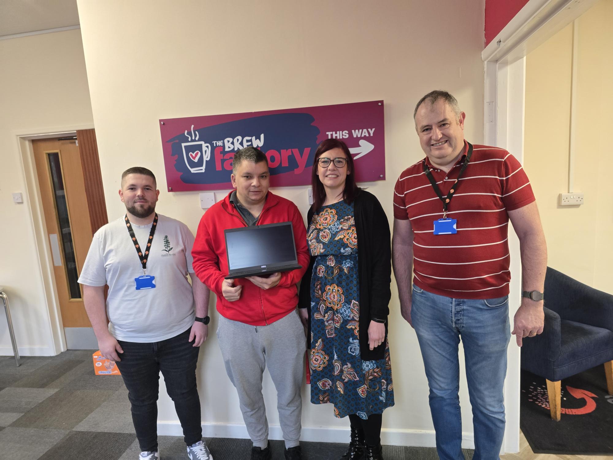 Trivallis Housing Landlord Wales Four people stand indoors in front of a sign that reads "The Brew Factory." One person holds a closed laptop. They are all wearing lanyards and looking at the camera.