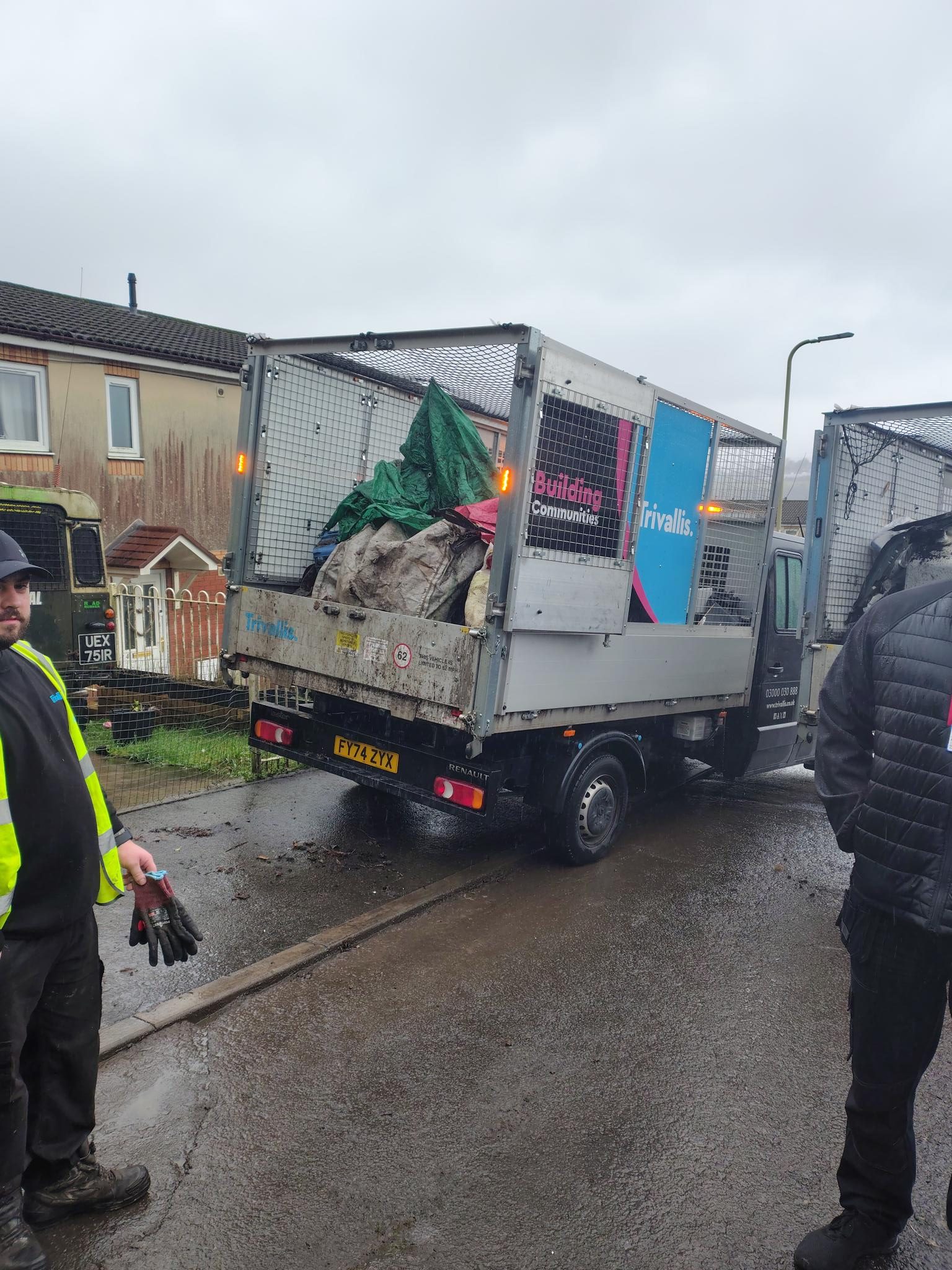 Trivallis Housing Landlord Wales A tipper truck with "Building Communities Trivallis" branding is parked on a wet residential street, loaded with various rubbish and covered with a green tarp. Two workers wearing high-visibility jackets stand nearby.