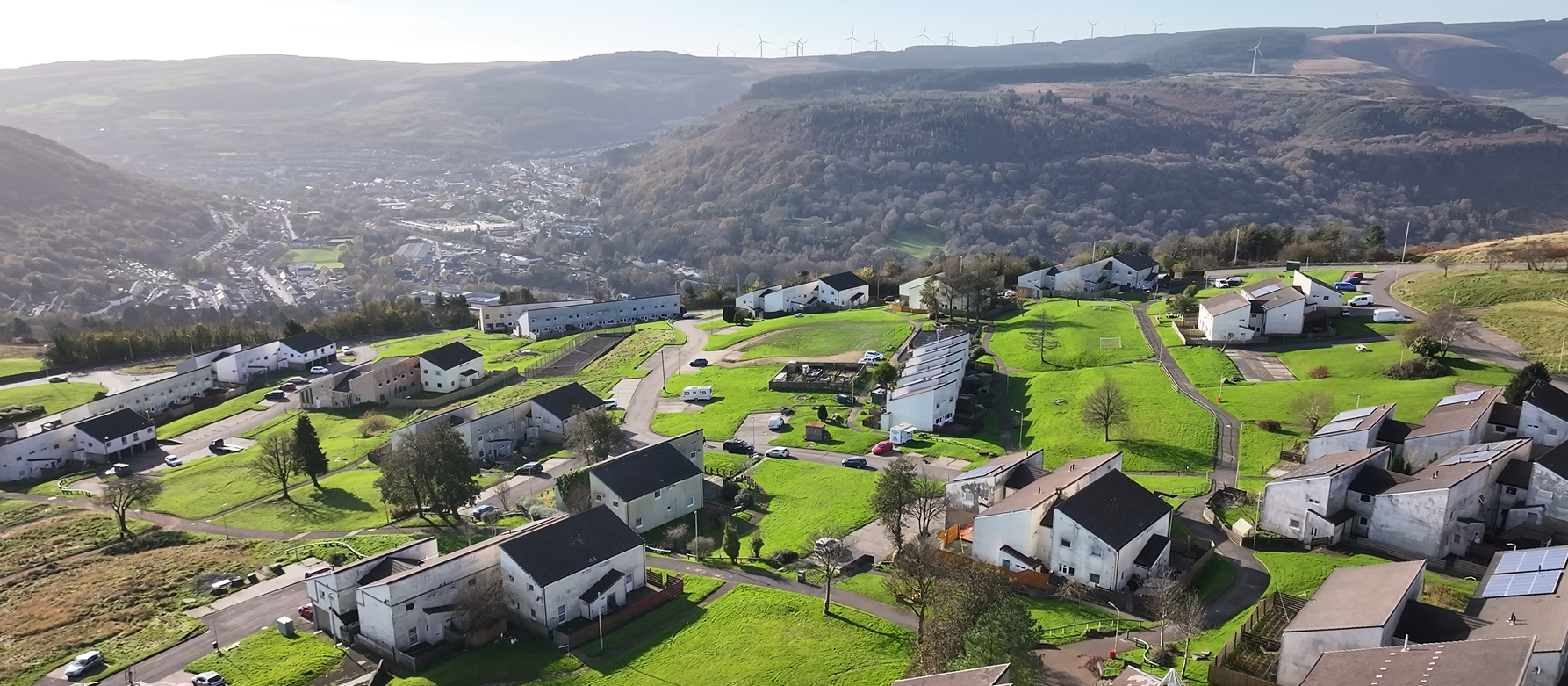 Trivallis Housing Landlord Wales Aerial view of a Penrhys with white houses and green lawns situated on hilly terrain, surrounded by forested hills. Wind turbines are visible on distant ridges under a clear sky.
