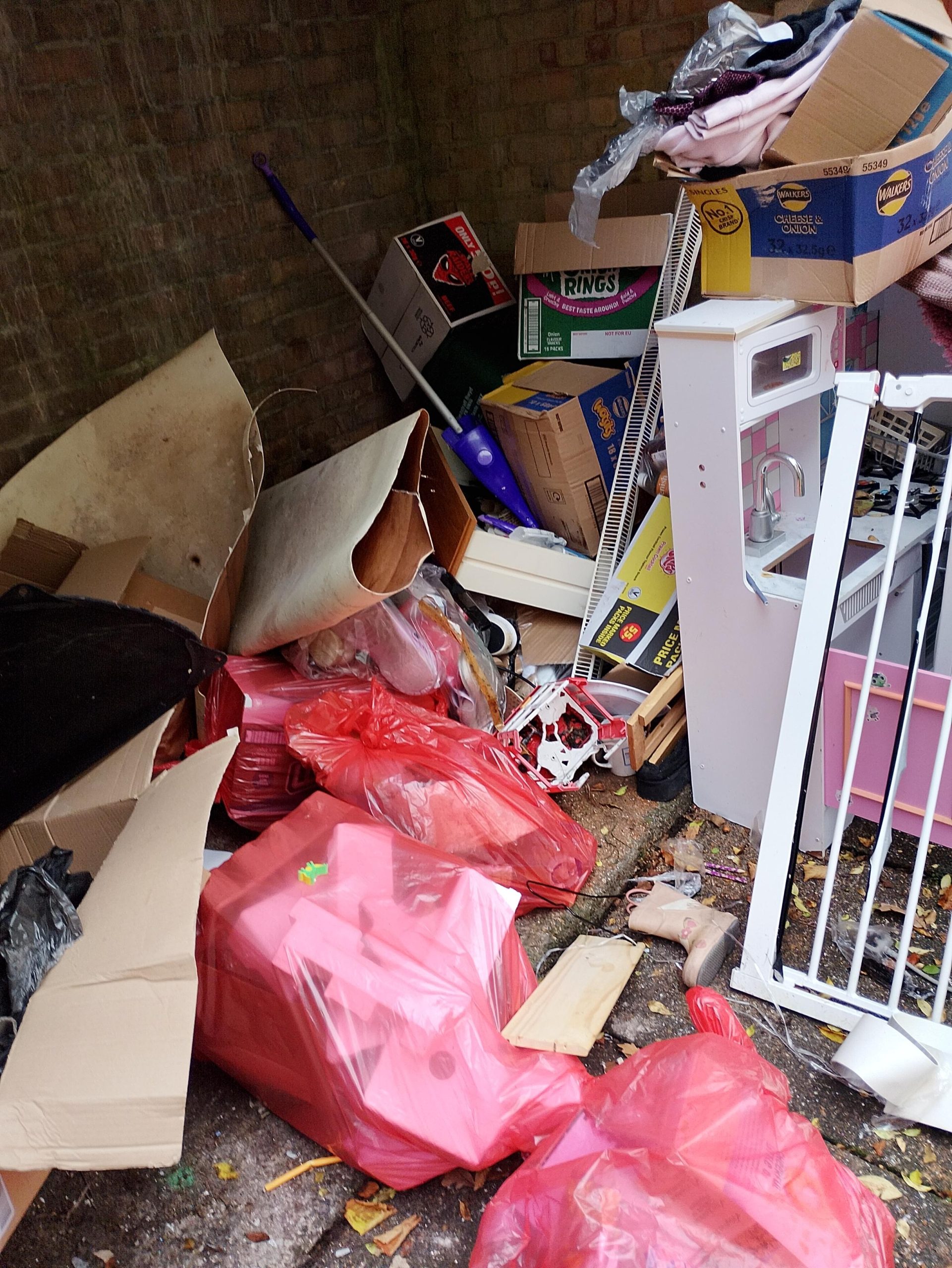 Trivallis Housing Landlord Wales A pile of assorted trash and discarded items, including red garbage bags, cardboard boxes, a mop, and broken furniture, is strewn in a corner against a brick wall.