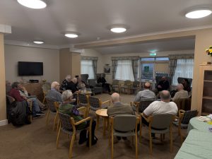 Trivallis Housing Landlord Wales A group of older adults sit in a circle of chairs in a well-lit community room, listening to a person who appears to be speaking at the front. The room has tables, a TV, and large windows with blinds.