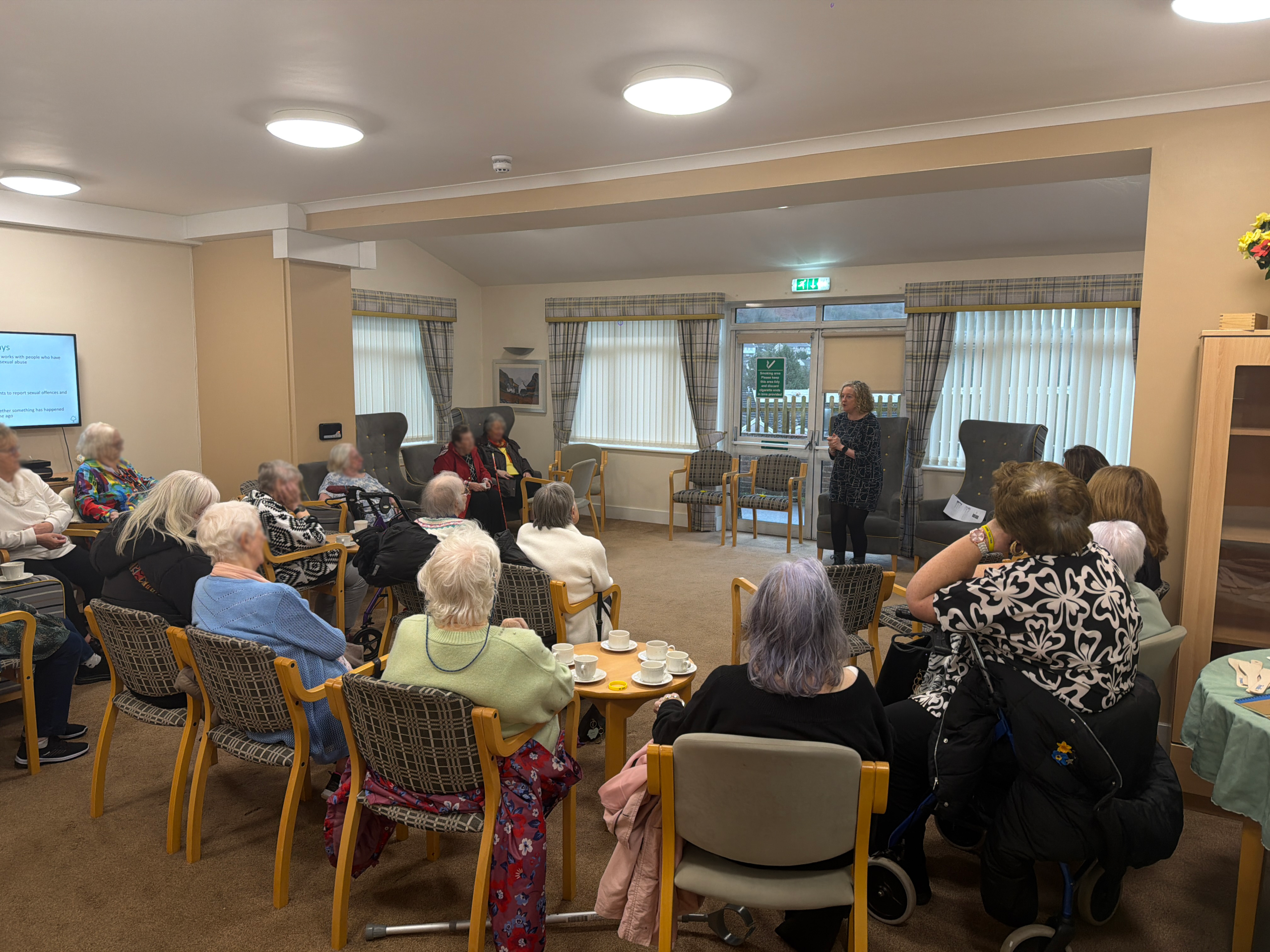Trivallis Housing Landlord Wales A group of elderly people sit in chairs around tables in a common room, watching a woman standing at the front speaking. There are cups and plates on the tables and large windows in the background.