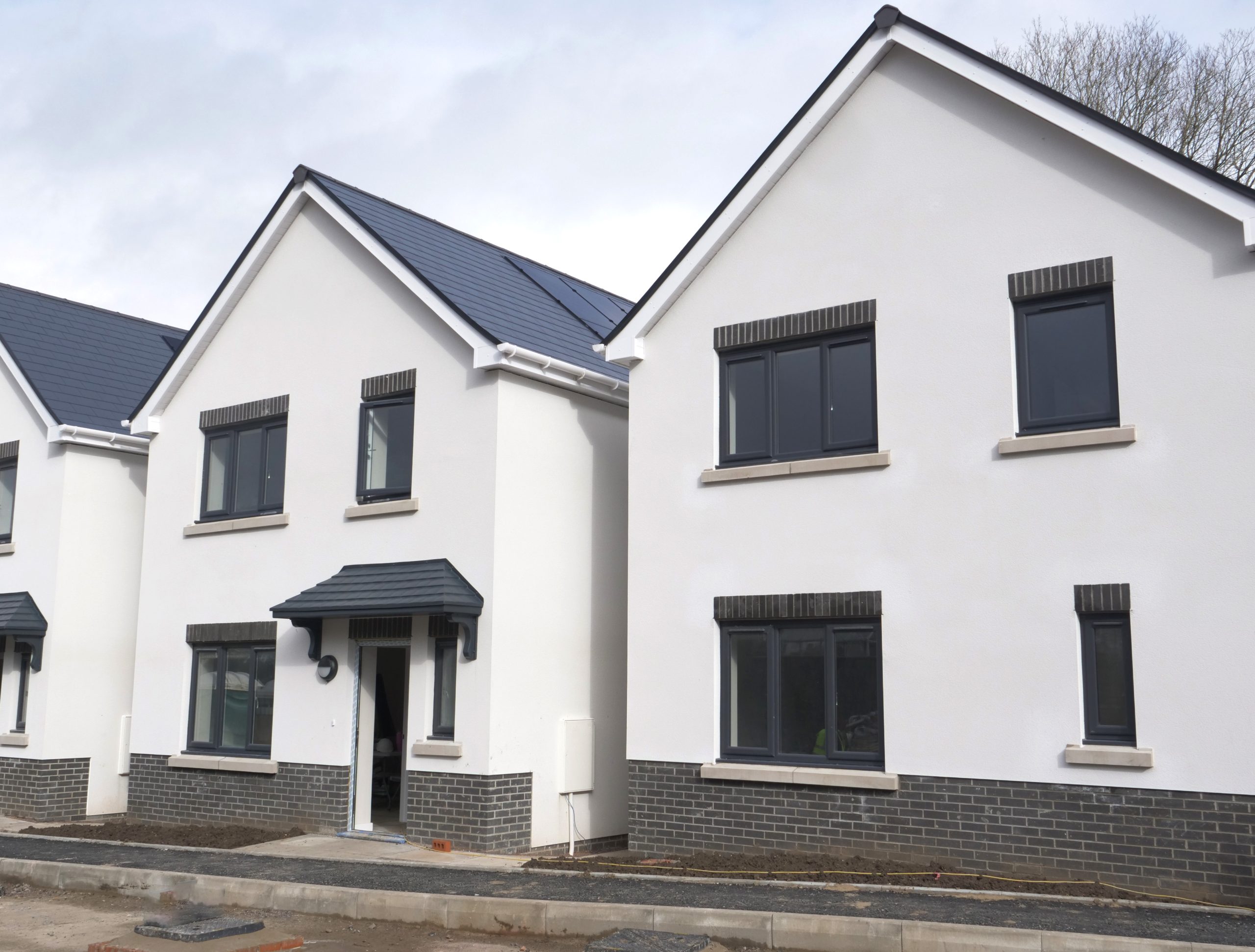 Trivallis Housing Landlord Wales Two newly built, modern detached houses with white walls, dark grey roof tiles, and brickwork at the base, situated on an unfinished street with no landscaping or finished pavement.