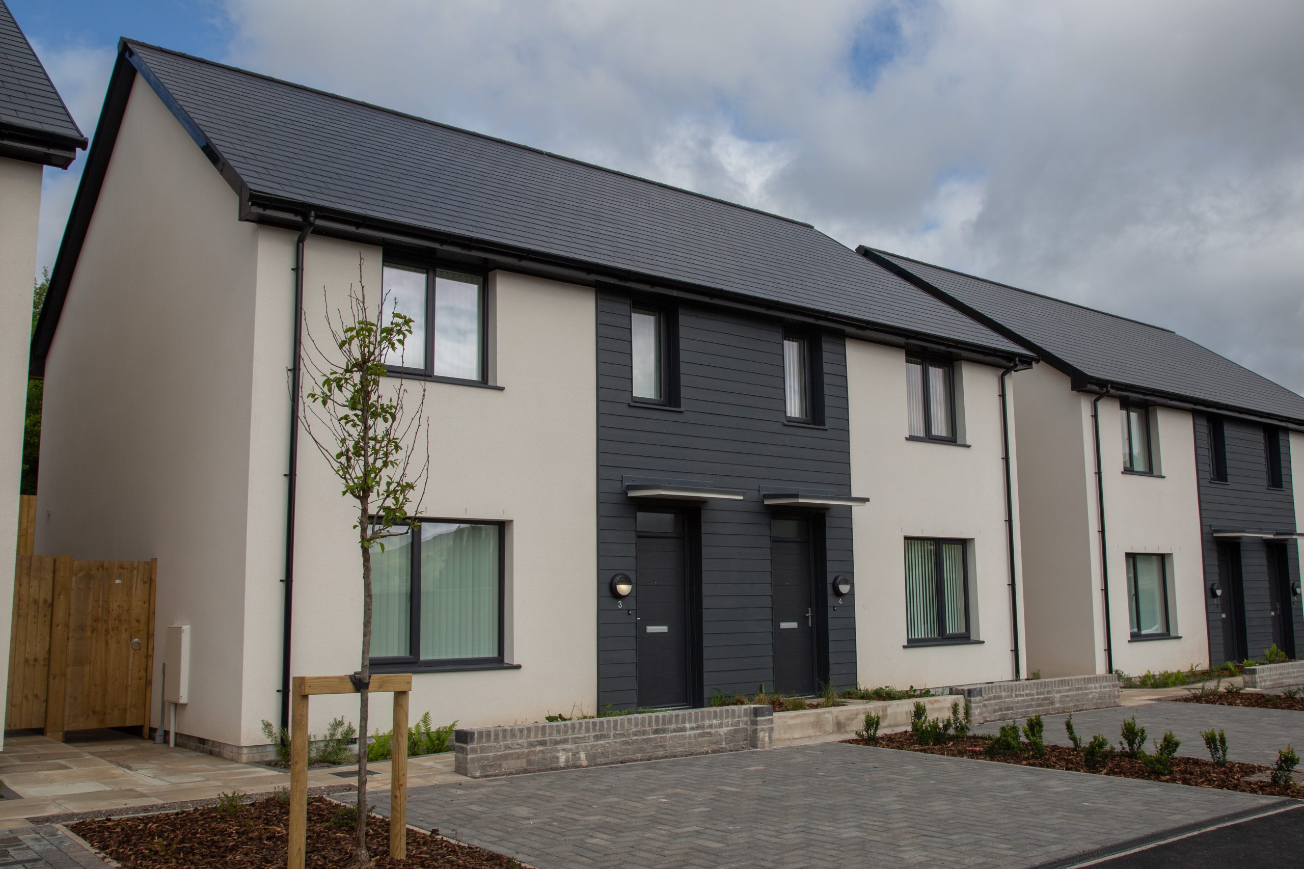 Trivallis Housing Landlord Wales Two modern, semi-detached houses with light-colored walls and dark grey accents. Each house has a small front garden, a young tree, and large front windows. The sky is partly cloudy.