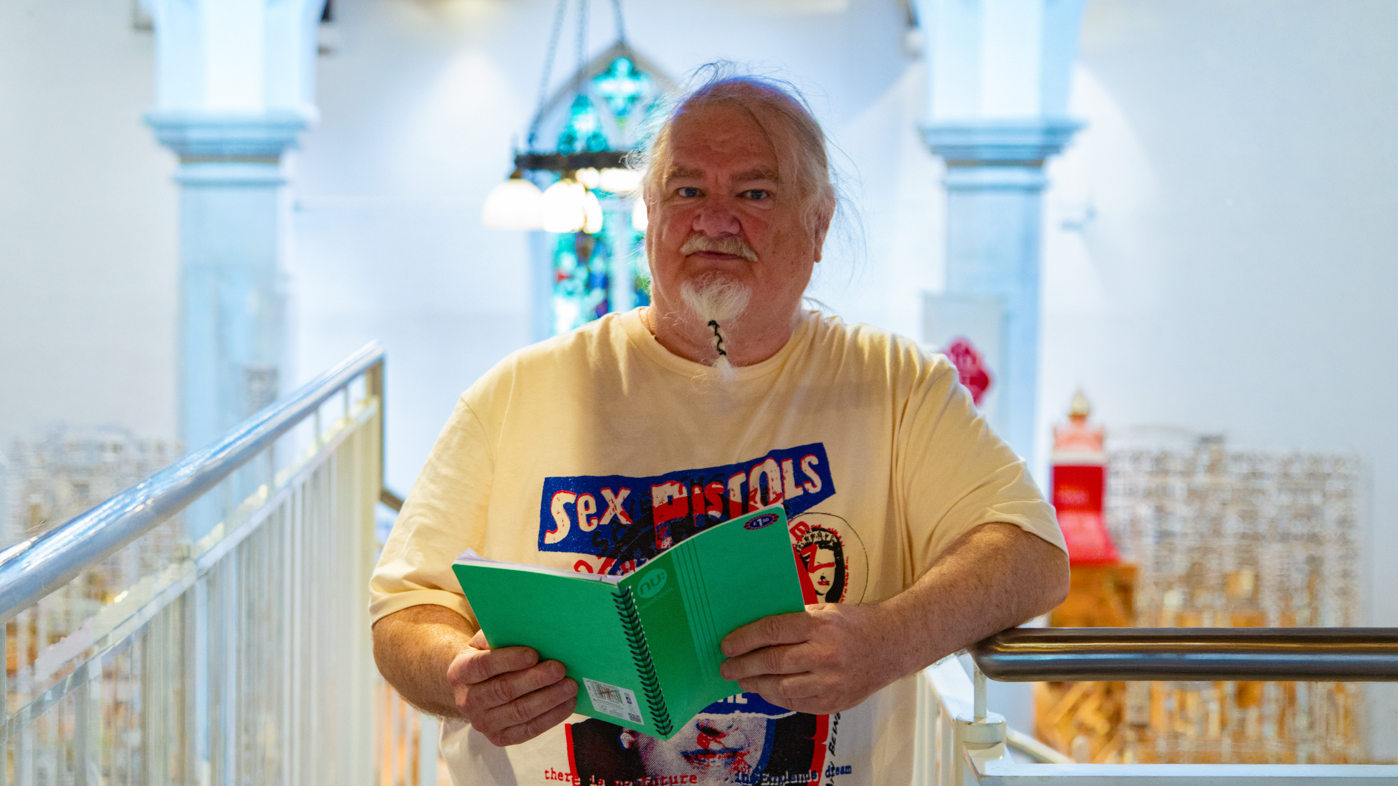 Trivallis Housing Landlord Wales An older man with long white hair and a beard stands indoors holding a green notebook. He wears a Sex Pistols T-shirt and stands in front of a balcony railing with columns, lights, and stained glass visible in the background.