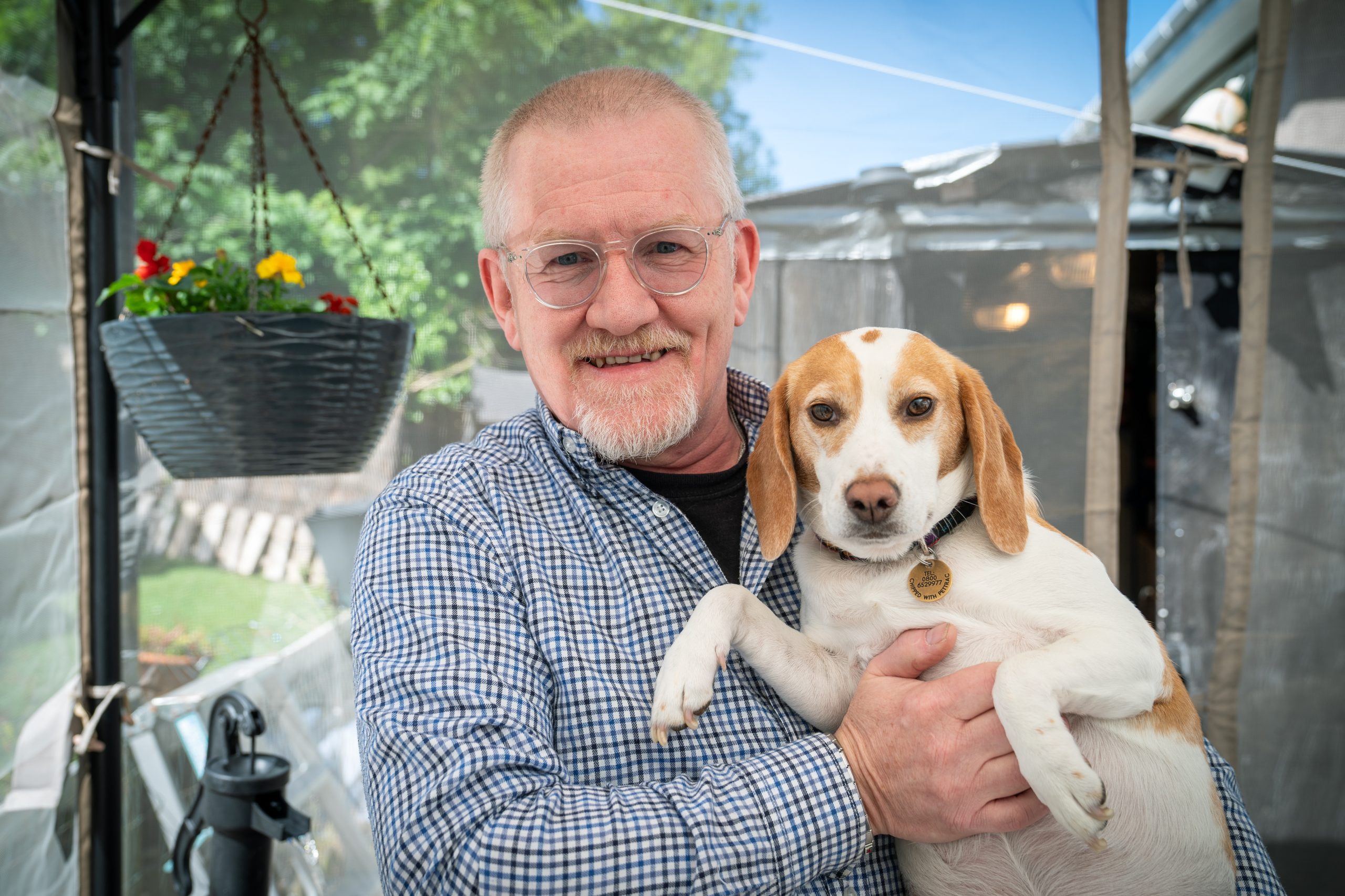 Trivallis Housing Landlord Wales An older man with glasses smiles while holding a beagle dog in his arms. They are outside near a hanging plant and a clear tent, with greenery and a wooden fence in the background.