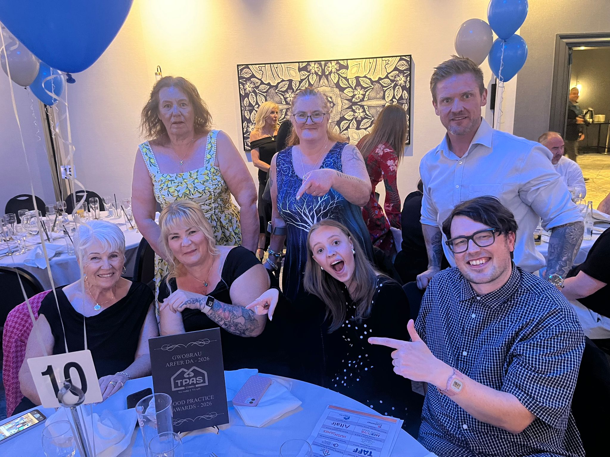 Trivallis Housing Landlord Wales Seven people are gathered around a round table at a formal event, some sitting, some standing, with a sign on the table and blue and white balloons nearby. Most are smiling and pointing at the sign.