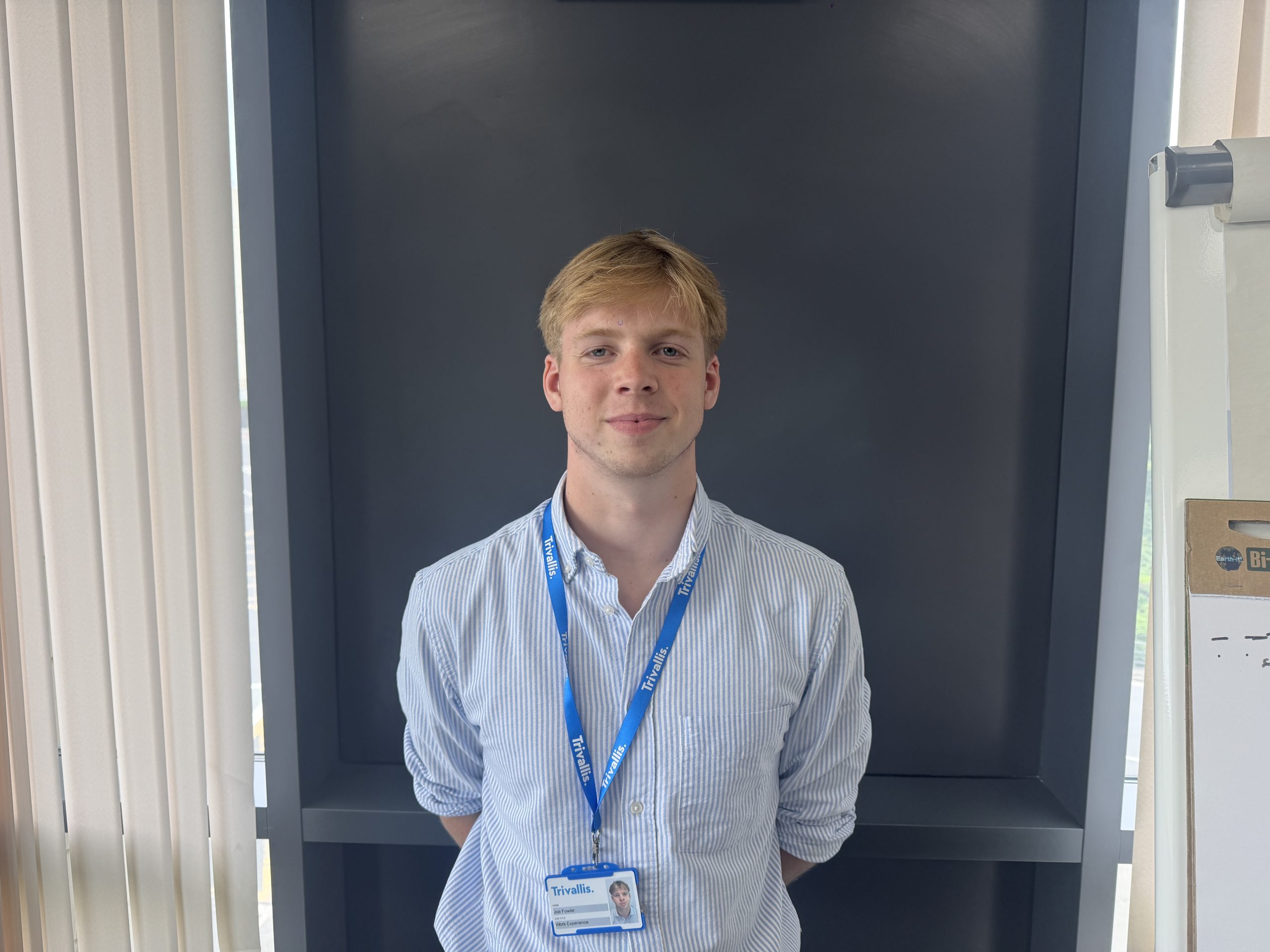 Trivallis Housing Landlord Wales A person with short blond hair, wearing a light blue striped shirt and a blue lanyard with an ID badge, stands in front of a dark gray background. Light comes from windows with vertical blinds on the sides.