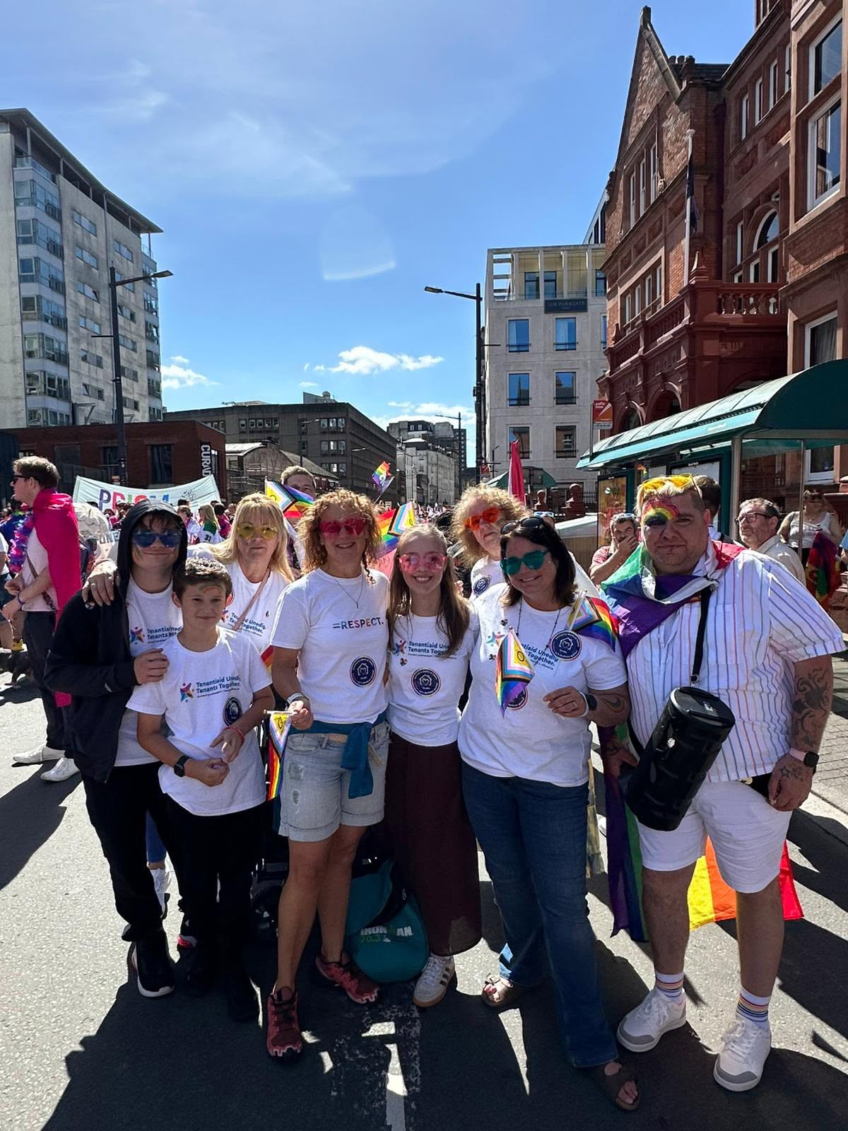 Trivallis Housing Landlord Wales A group of people wearing matching white shirts with logos pose together on a sunny city street during a parade, holding rainbow flags and accessories. There are buildings and other parade participants in the background.