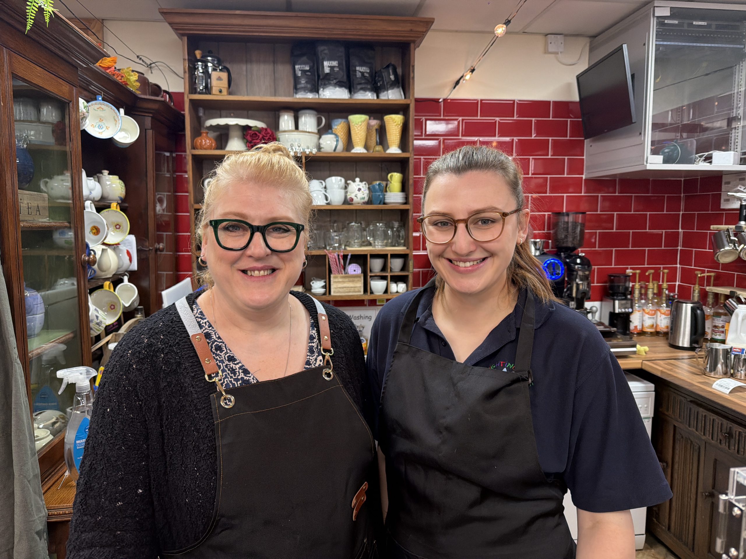 Trivallis Housing Landlord Wales Two women wearing black aprons stand side by side and smile in a café setting, with red tile walls, shelves of cups, jars, and coffee equipment visible in the background.