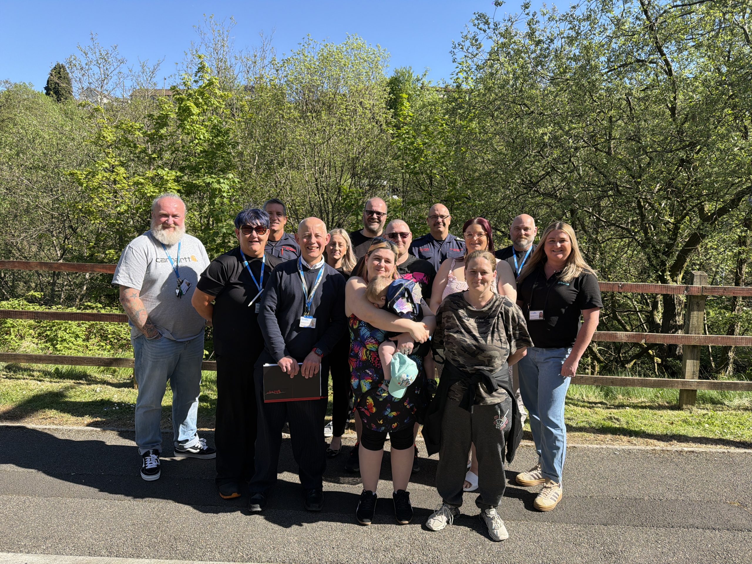 Trivallis Housing Landlord Wales A group of fifteen adults stands on a paved path in front of a wooden fence with trees and greenery behind them on a sunny day. Some people are smiling, and most are wearing casual clothing and lanyards.