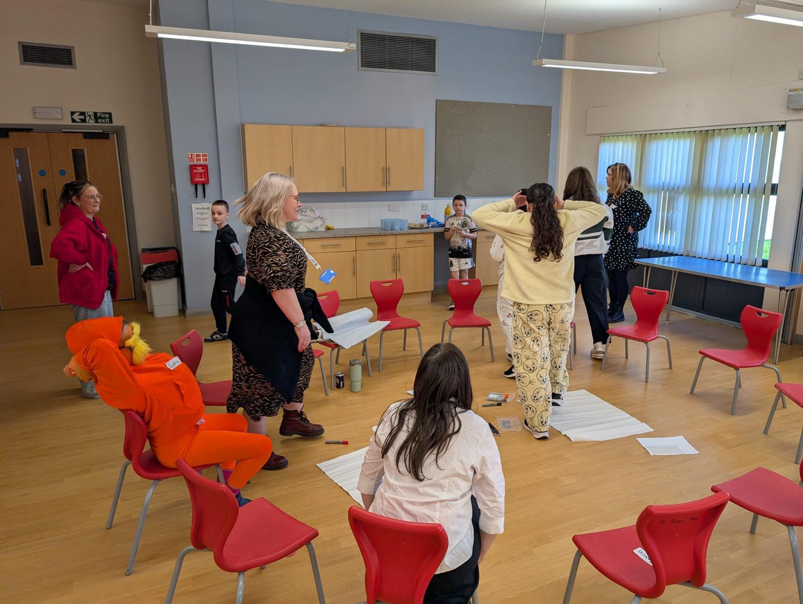 Trivallis Housing Landlord Wales A group of people are in a room with red chairs arranged in a circle. Some stand, some sit, and a few appear to be holding papers. The room has wooden floors, large windows, and kitchen cabinets in the background.