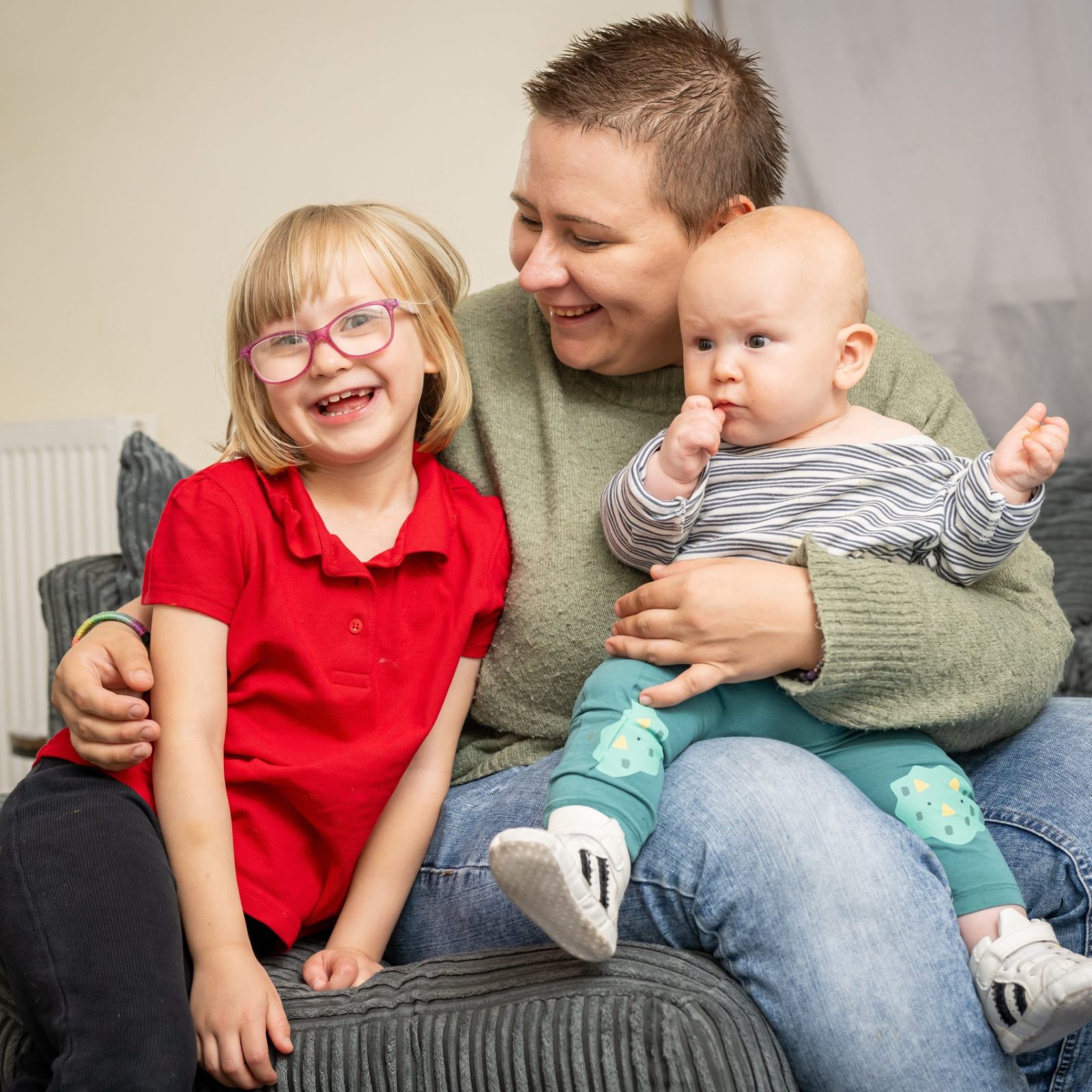 Trivallis Housing Landlord Wales A woman sitting on a sofa holds a baby in their lap and wraps an arm around a smiling young girl with glasses and a red shirt. They are indoors, in a living room with gray furniture.
