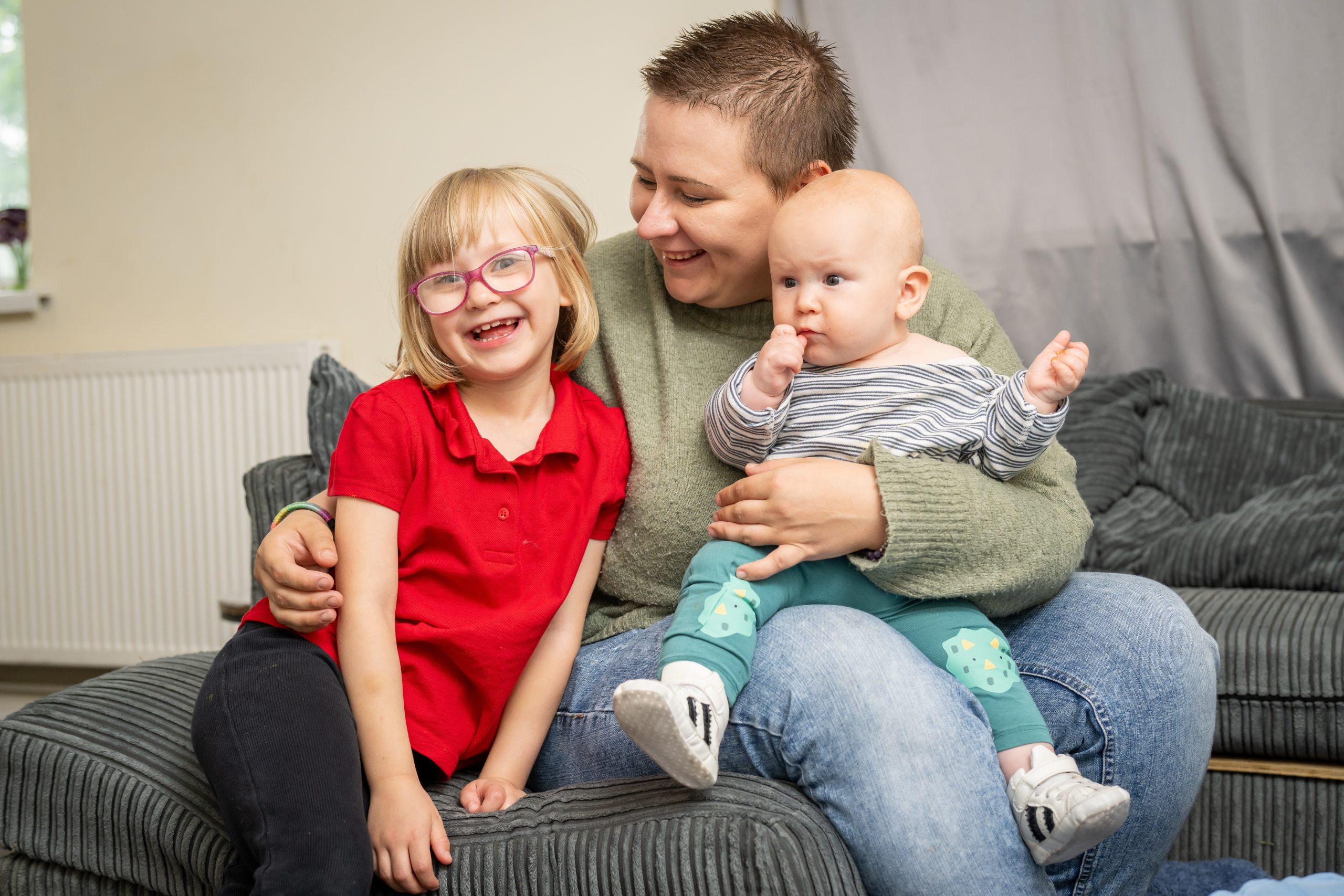 Trivallis Housing Landlord Wales A woman sitting on a sofa holds a baby in their lap and wraps an arm around a smiling young girl with glasses and a red shirt. They are indoors, in a living room with gray furniture.