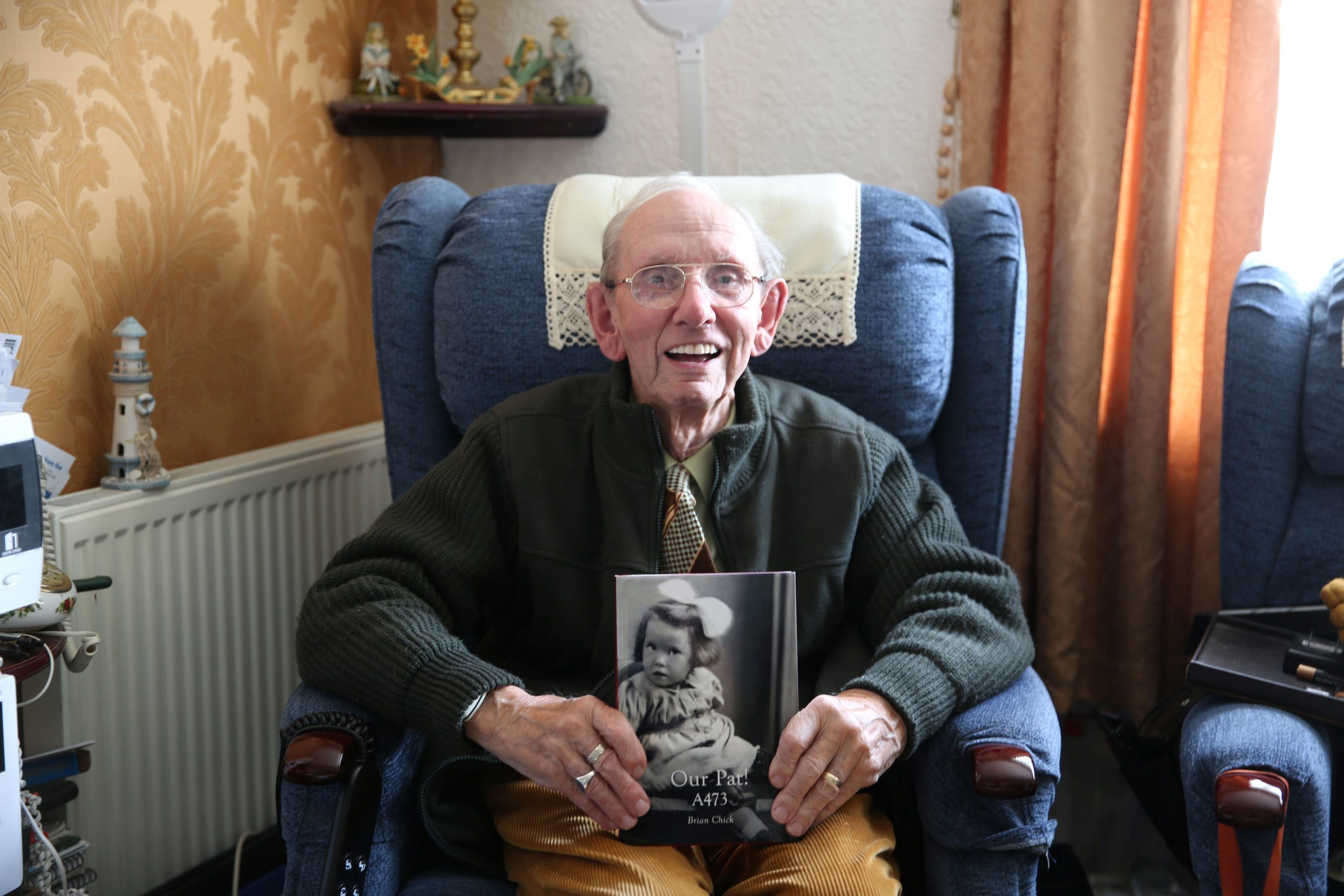 Trivallis Housing Landlord Wales An elderly man sits in a blue armchair, smiling and holding an open book with a black-and-white photo of a child on the cover. Figurines and a lamp are visible on a shelf and table in the background.