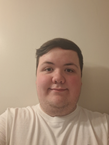 Trivallis Housing Landlord Wales A person with short dark hair and a trimmed beard is wearing a white shirt and standing in front of a plain light-colored wall, looking directly at the camera with a neutral expression.