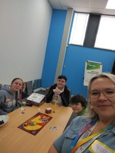 Trivallis Housing Landlord Wales Four people sit around a table playing a board game in a bright room. The game pieces and cards are on the table, and one person is taking a selfie. A flip chart and a poster are visible in the background.