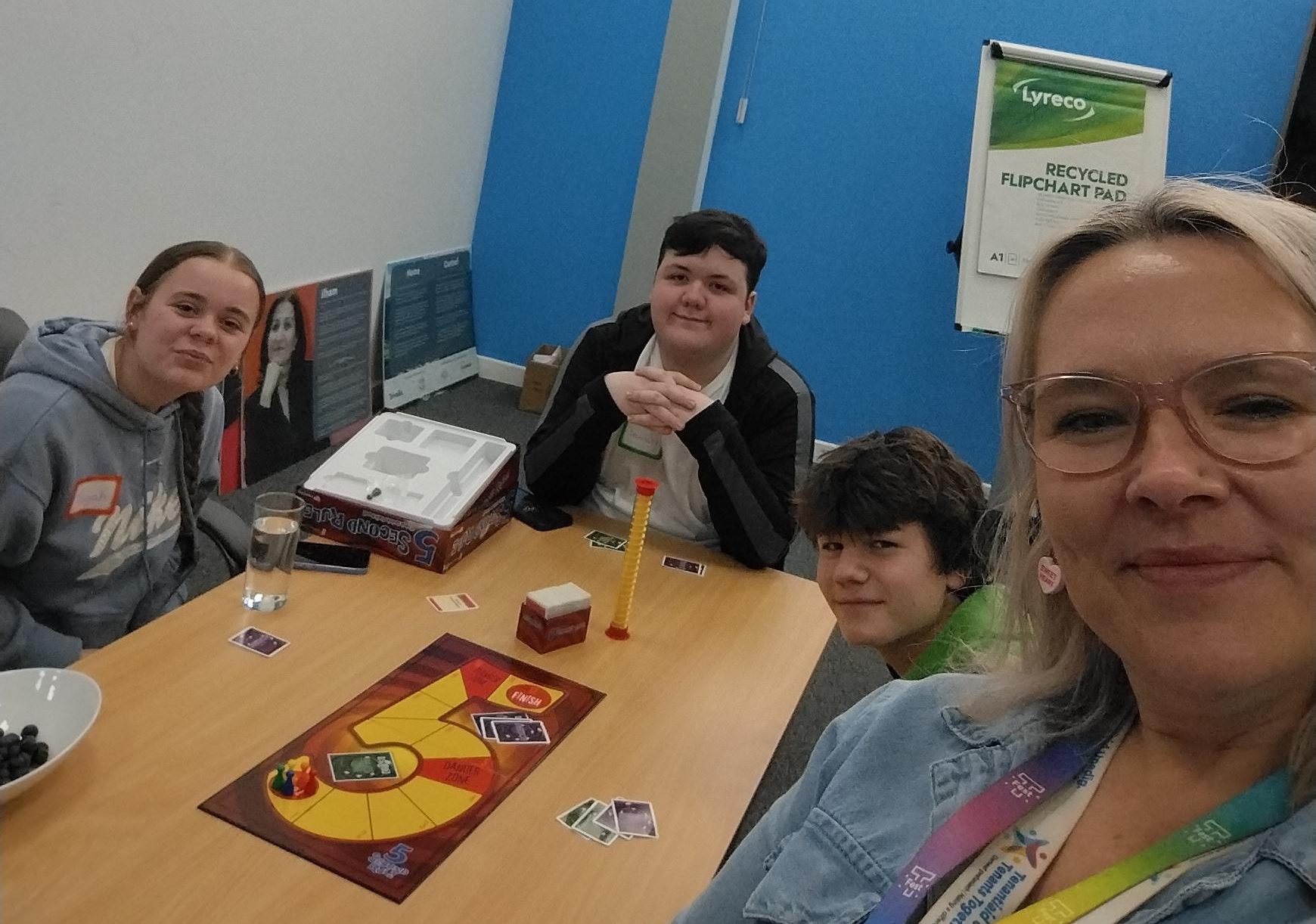 Trivallis Housing Landlord Wales Four people sit around a table playing a board game in a bright room. The game pieces and cards are on the table, and one person is taking a selfie. A flip chart and a poster are visible in the background.