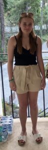 Trivallis Housing Landlord Wales A young woman with long brown hair stands on a balcony, wearing a black tank top, beige high-waisted shorts, and light sandals. She has sunglasses on her head. Bottled water is stacked to her left. Trees are visible in the background.