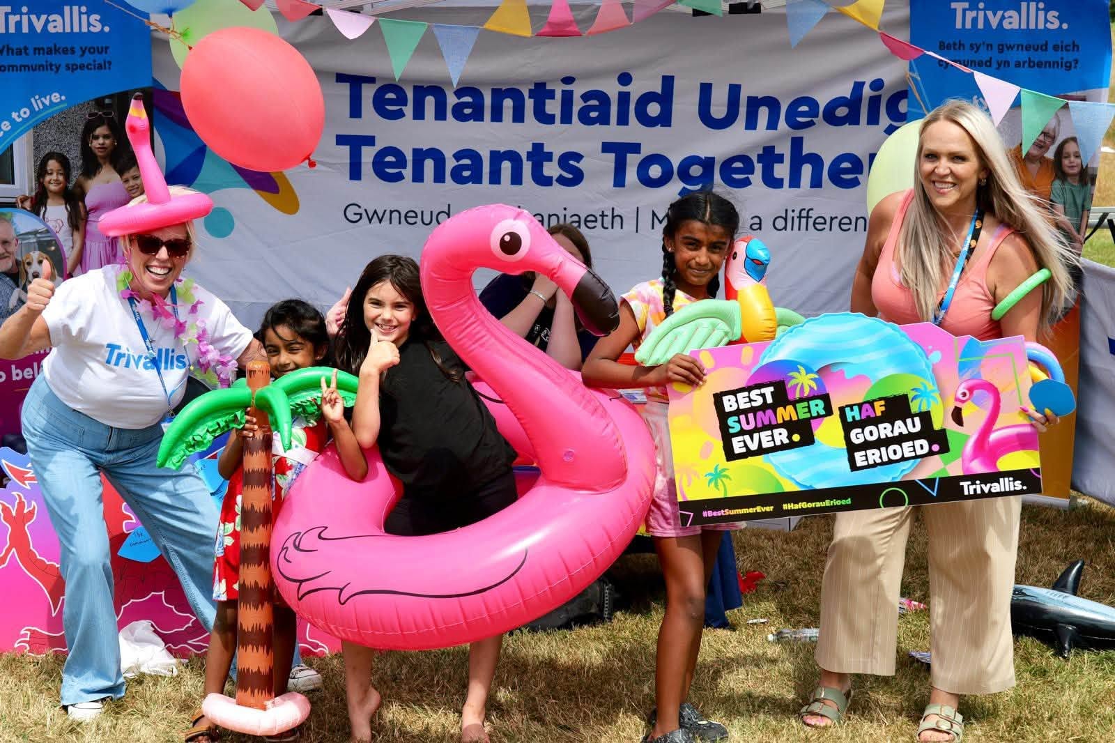 Trivallis Housing Landlord Wales Five people, including three children, pose outdoors with colorful inflatable toys like a flamingo and palm tree, in front of a banner reading “Tenants Together” and “Trivallis” at a community event.