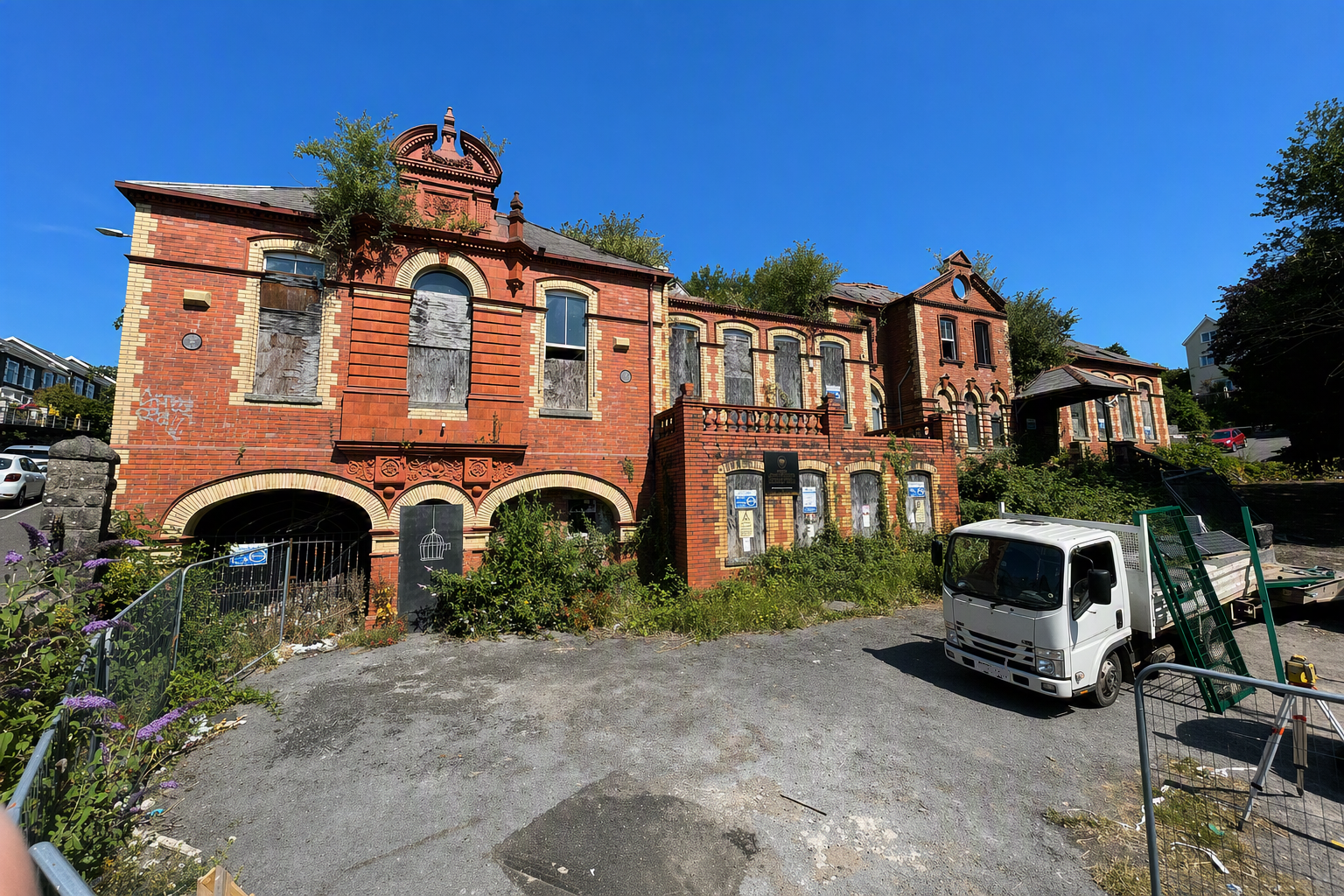 Trivallis Housing Landlord Wales Merthyr General Hospital, an old, abandoned red-brick building with boarded-up windows and overgrown plants is pictured under a clear blue sky. A white truck and construction materials are visible in the foreground.
