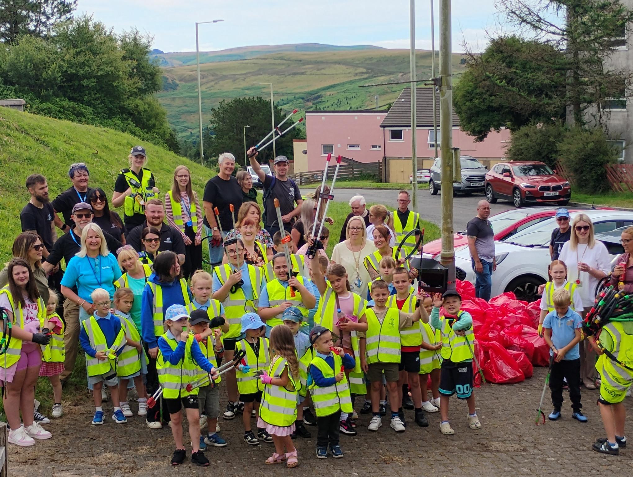 Trivallis Housing Landlord Wales A group of adults and children in yellow safety vests stand outdoors with litter pickers and full red trash bags. They pose for a photo in a residential area with green hills in the background.