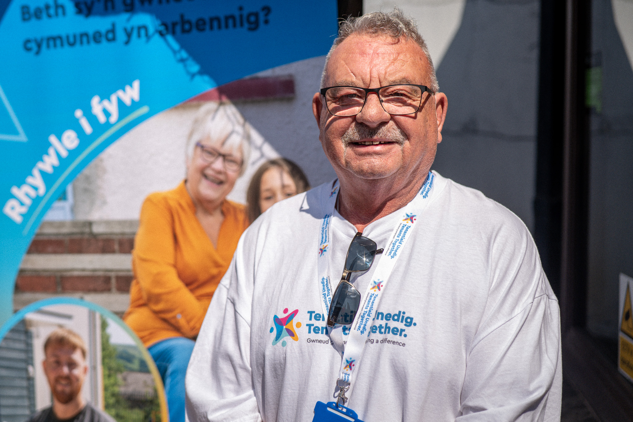 Trivallis Housing Landlord Wales An older man wearing glasses and a “Ten for a Difference” lanyard and T-shirt stands in front of a blue promotional banner with Welsh and English text, smiling at the camera in bright sunlight.