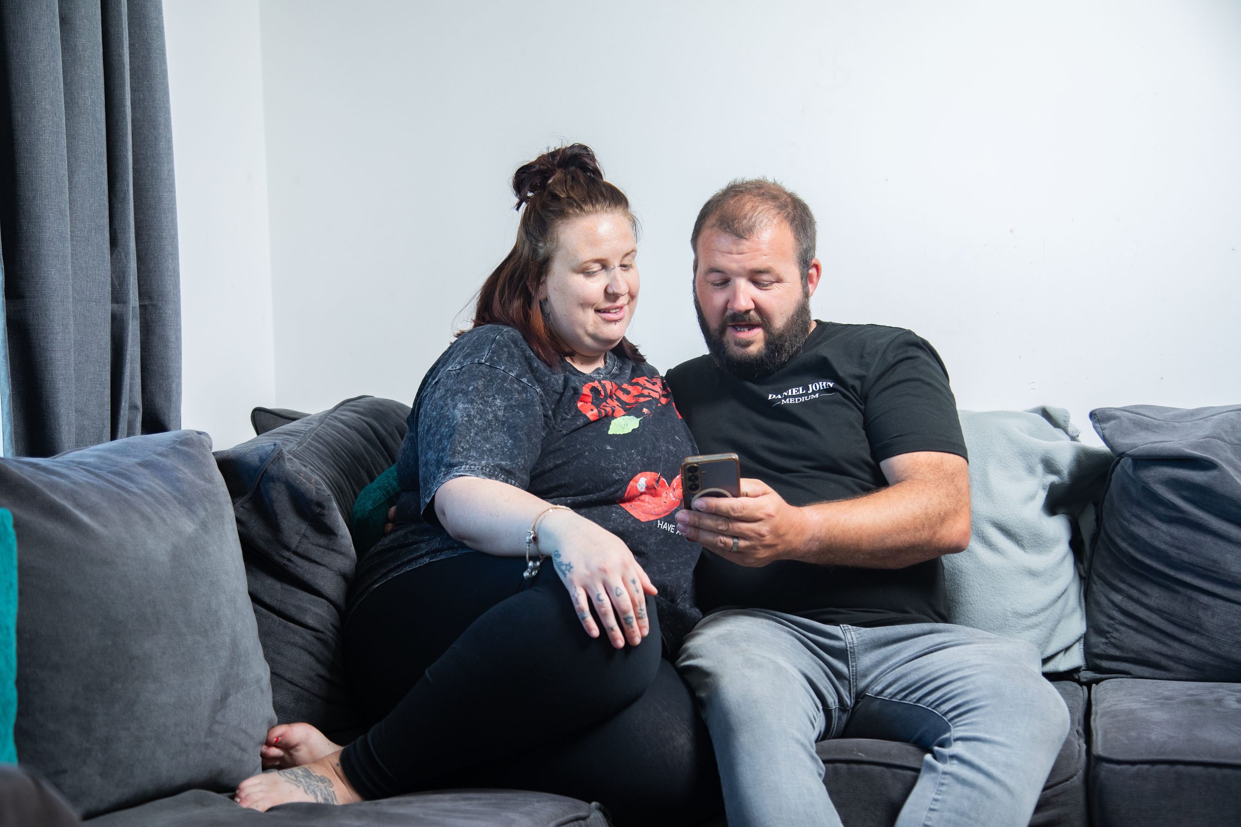 Trivallis Housing Landlord Wales A woman and a man are sitting together on a grey sofa, looking at a mobile phone that the man is holding. They appear to be sharing or discussing something on the screen.