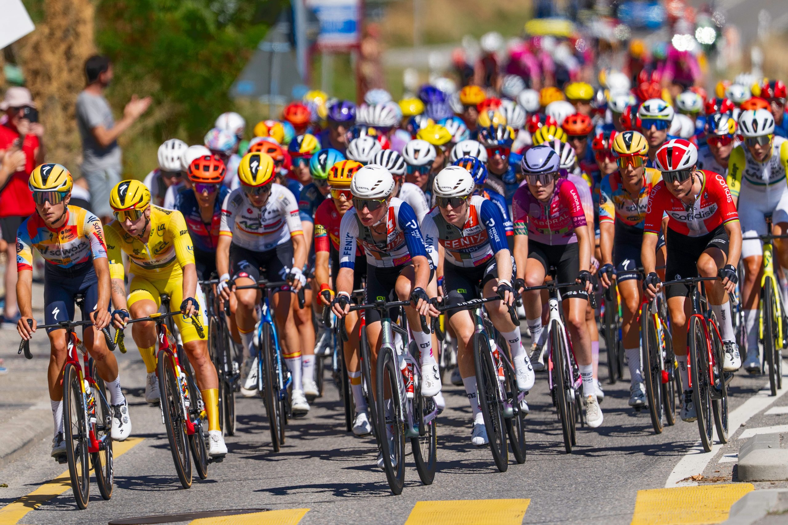 Trivallis Housing Landlord Wales A large group of cyclists wearing colourful jerseys and helmets rides closely together during a road race on a sunny day. Spectators and trees line the road in the background.
