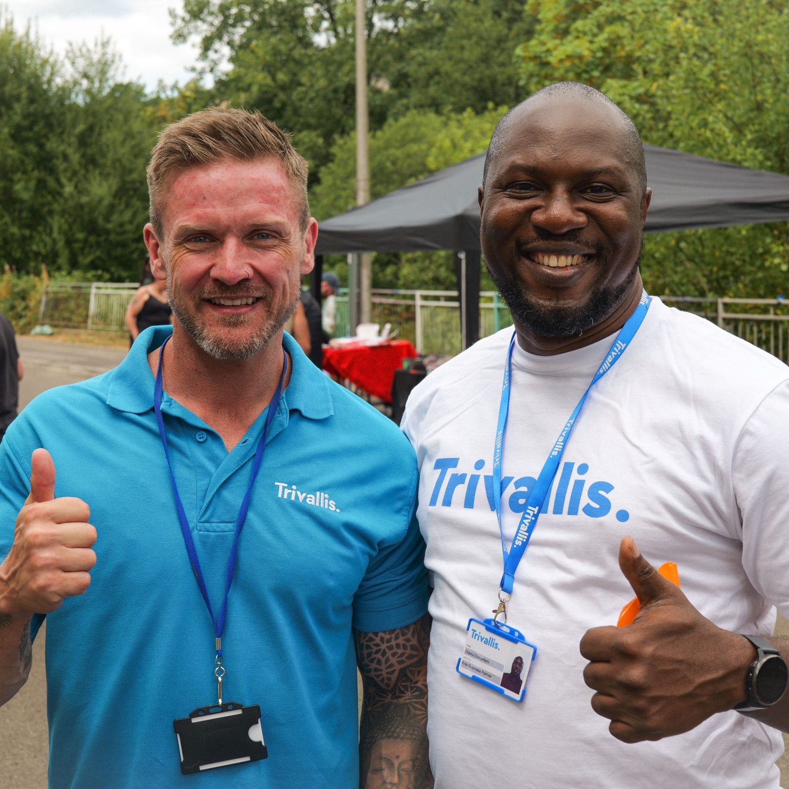 Trivallis Housing Landlord Wales Two men wearing Trivallis branded shirts and lanyards smile and give thumbs-up gestures outdoors.